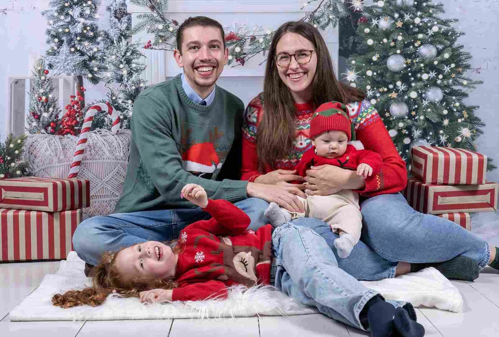  Older sister holding her baby sibling on a white blanket in front of a Christmas tree and fireplace set. 