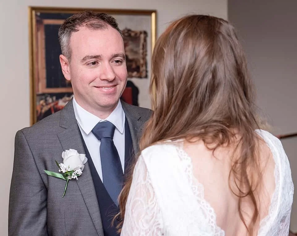 Joyful moments from a Dublin Registry Office wedding. A groom in a grey suit with a white rose boutonniere and a bride in a lace dress share a heartfelt look during their ceremony. The groom signs the official marriage documents, capturing the essenc