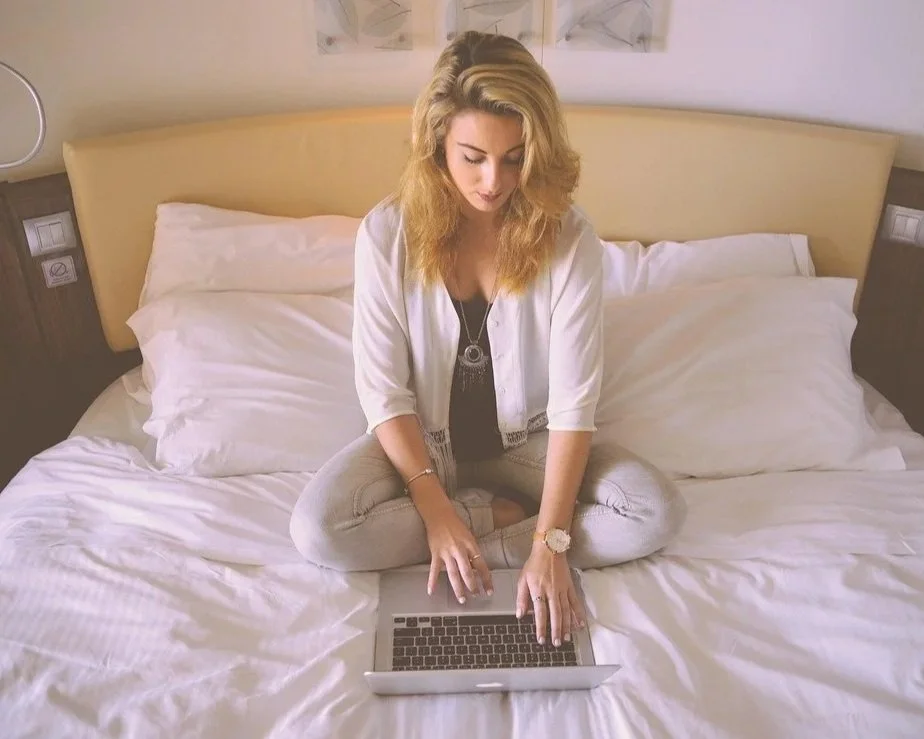 A white femme presenting person therapy client sitting on a bed, dressed casually, and. working on a laptop.