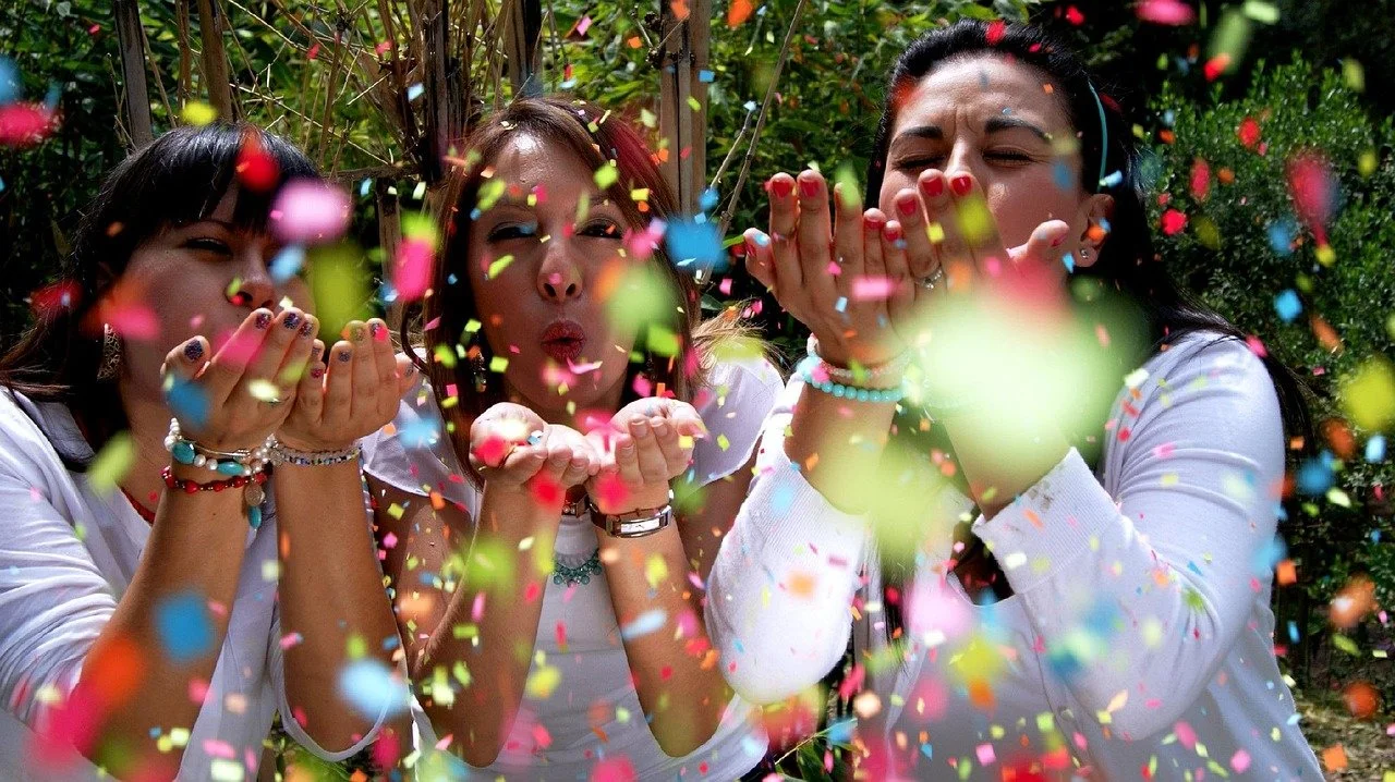 Three women surrounded by colorful confetti, blowing kisses and smiling outdoors.