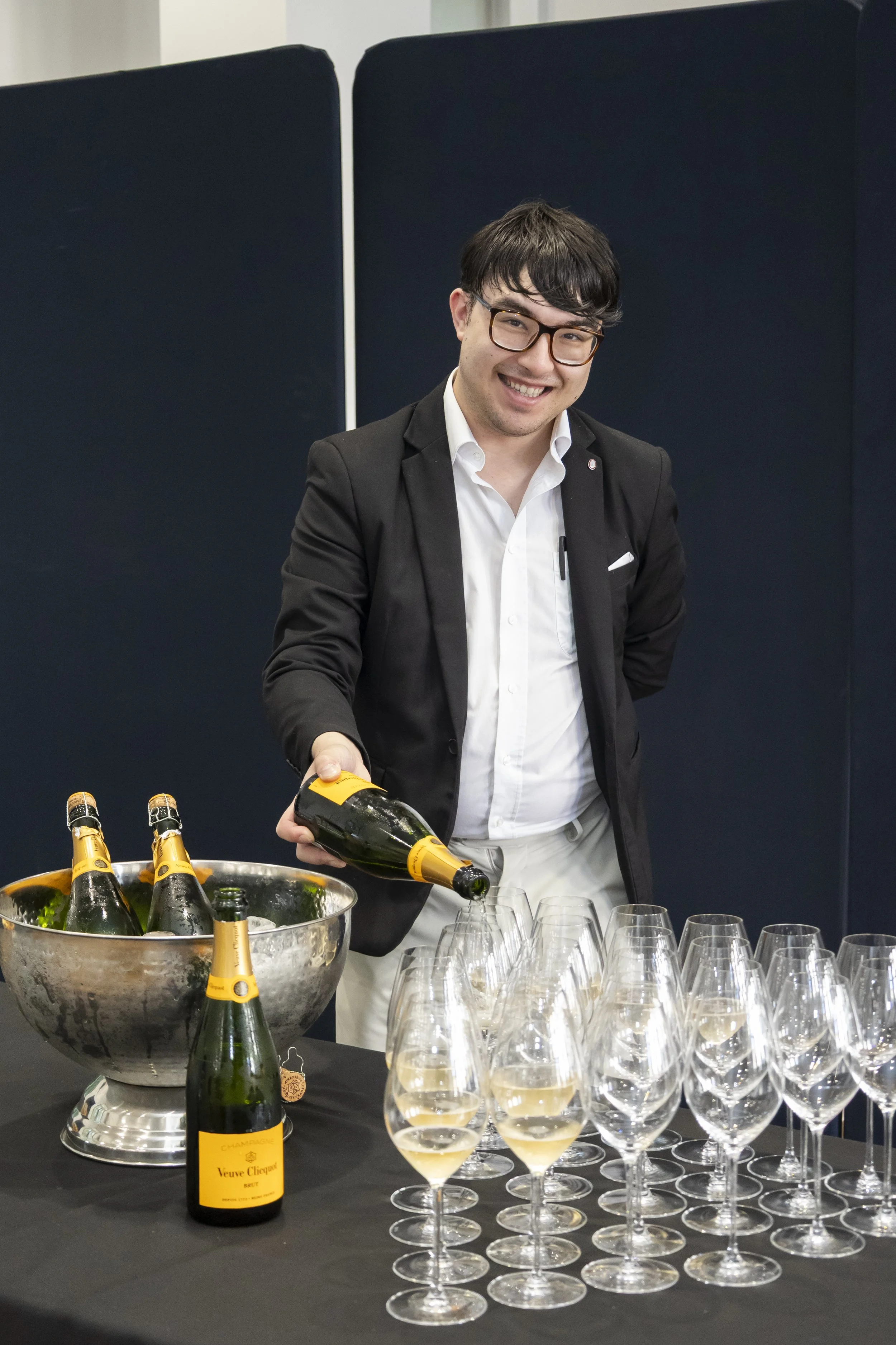 A well-dressed and jacketed smiling Brian, pouring champagne sparkling wine into glassware, accompanied by a cold ice bucket of various un opened bottles of Veuve Clicquot.