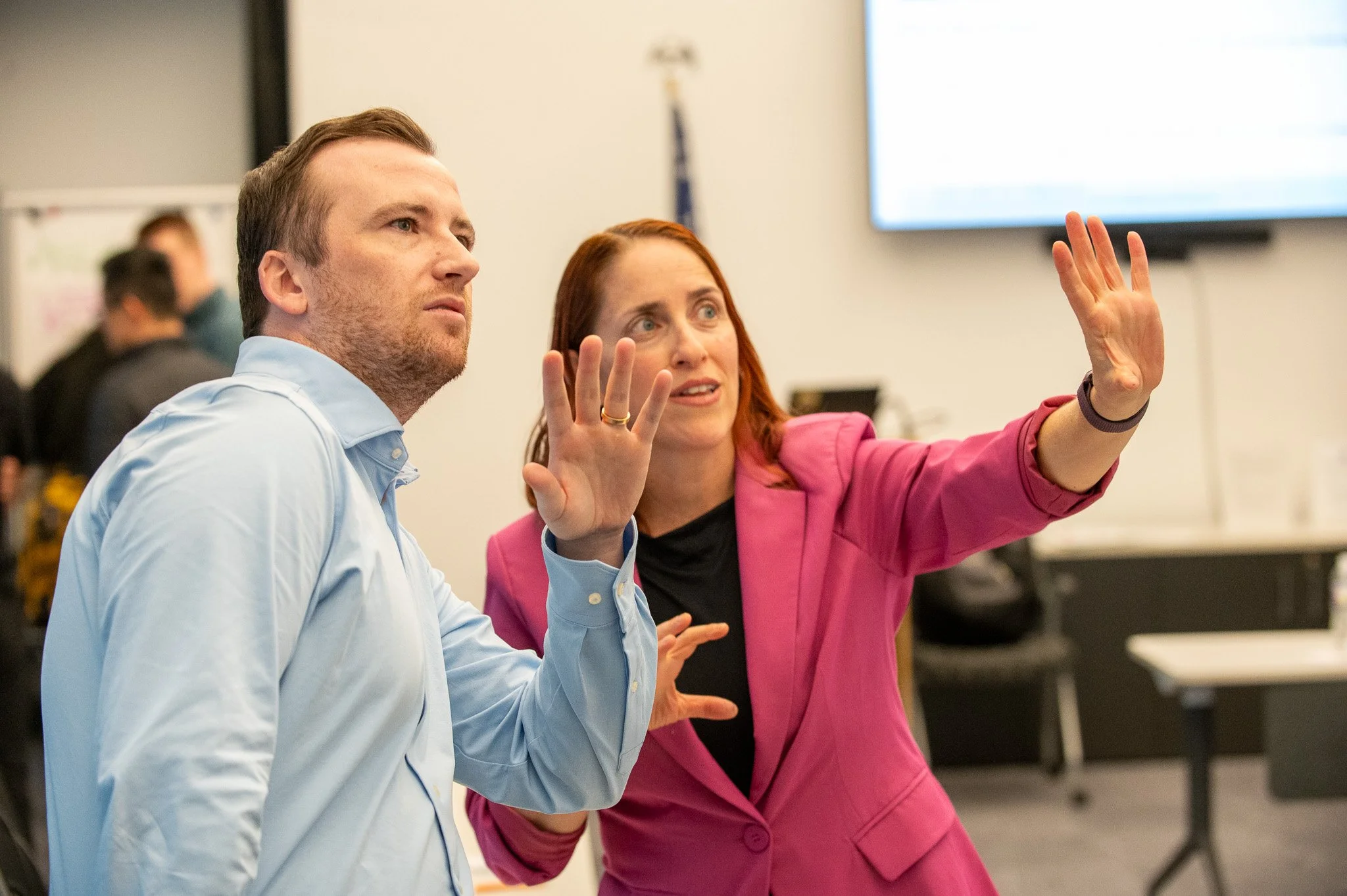 A woman in a pink blazer and a man in a light blue shirt are engaged in a discussion in a professional setting, with the woman gesturing with her hand.