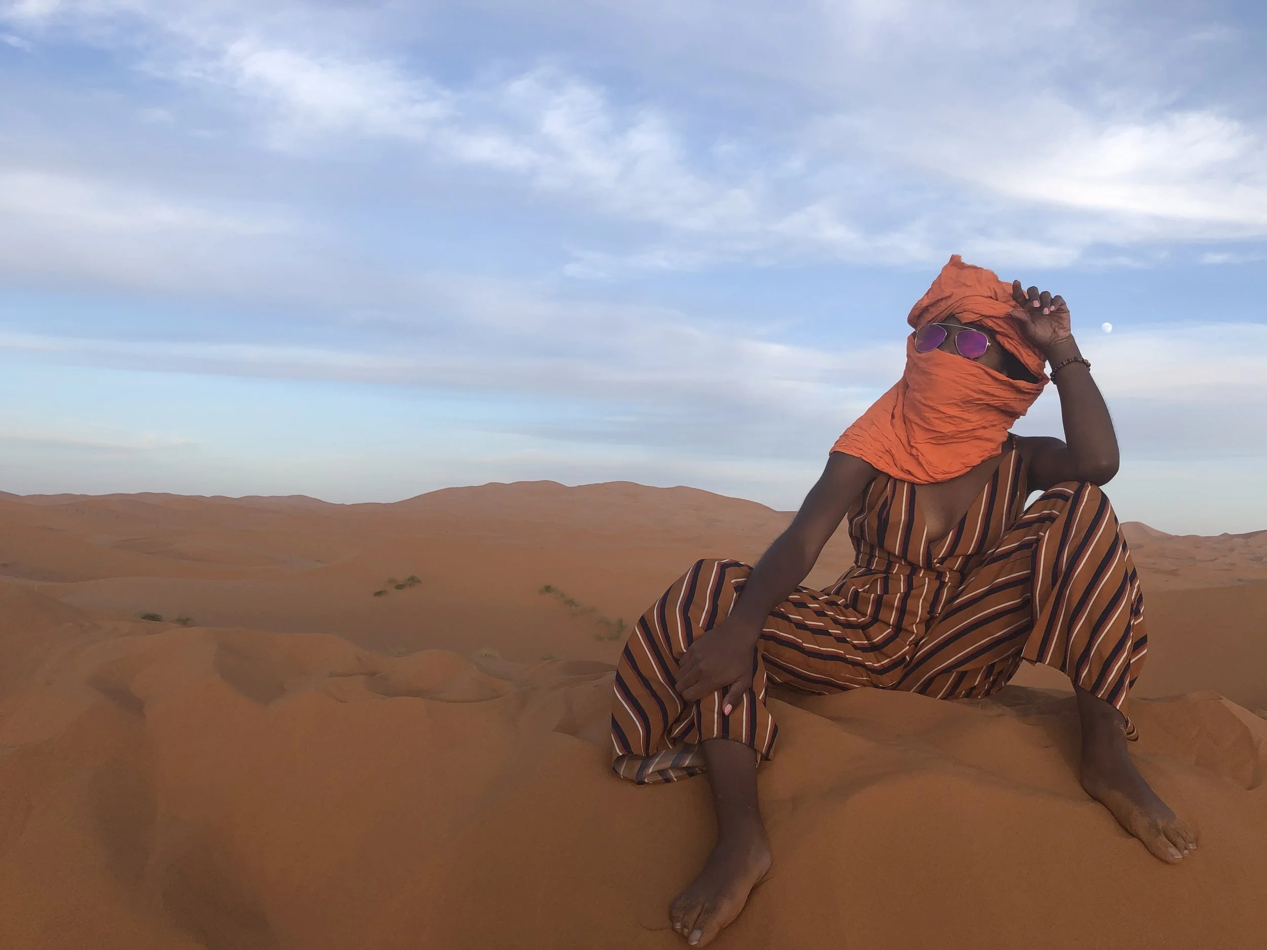 A young woman (Zephaniah) in the Sahara Desert wearing a striped outfit and head covering, sitting on sand dunes in a desert landscape under a partly cloudy sky with a small moon visible.