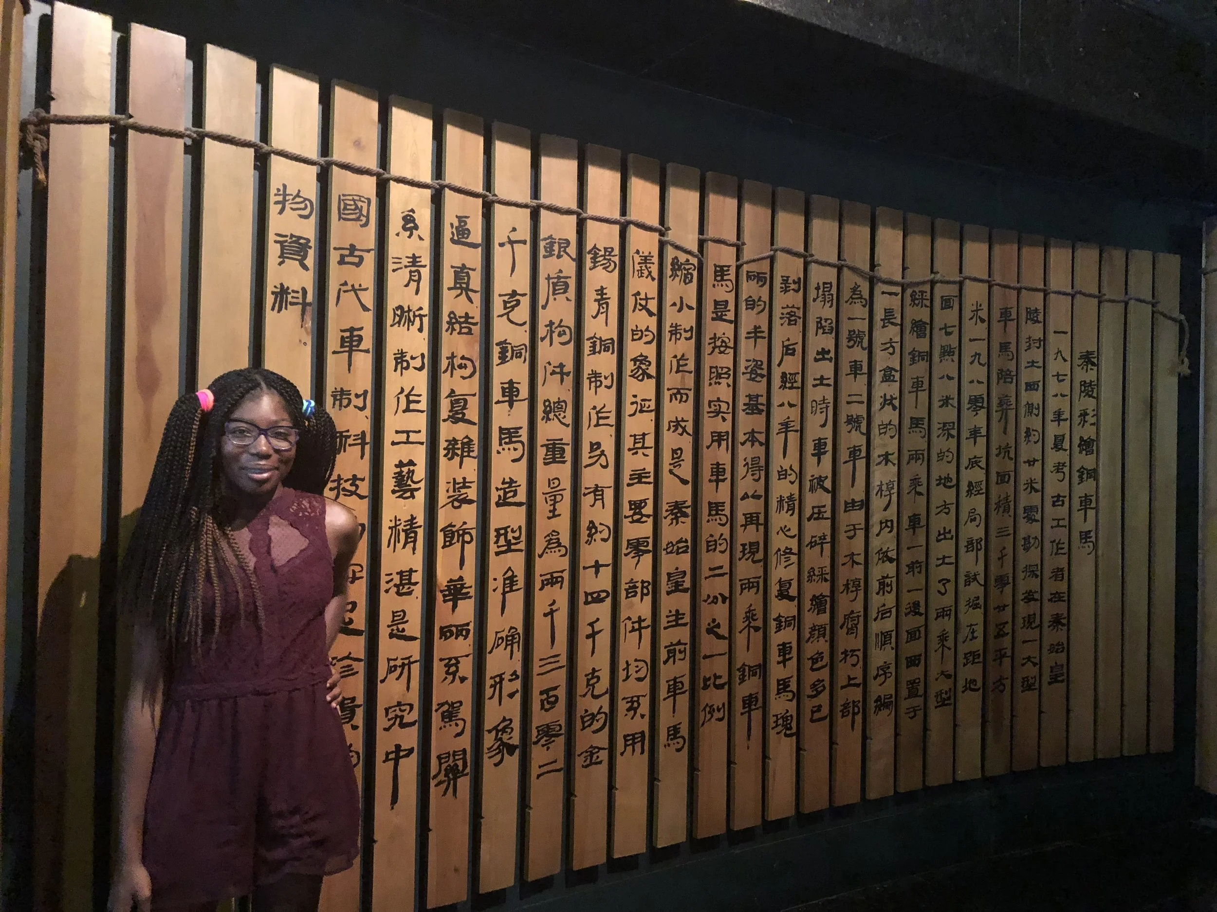 A young woman (Zephaniah) with braids, glasses, and a sleeveless maroon dress standing in front of a wooden wall with Chinese characters written on it.
