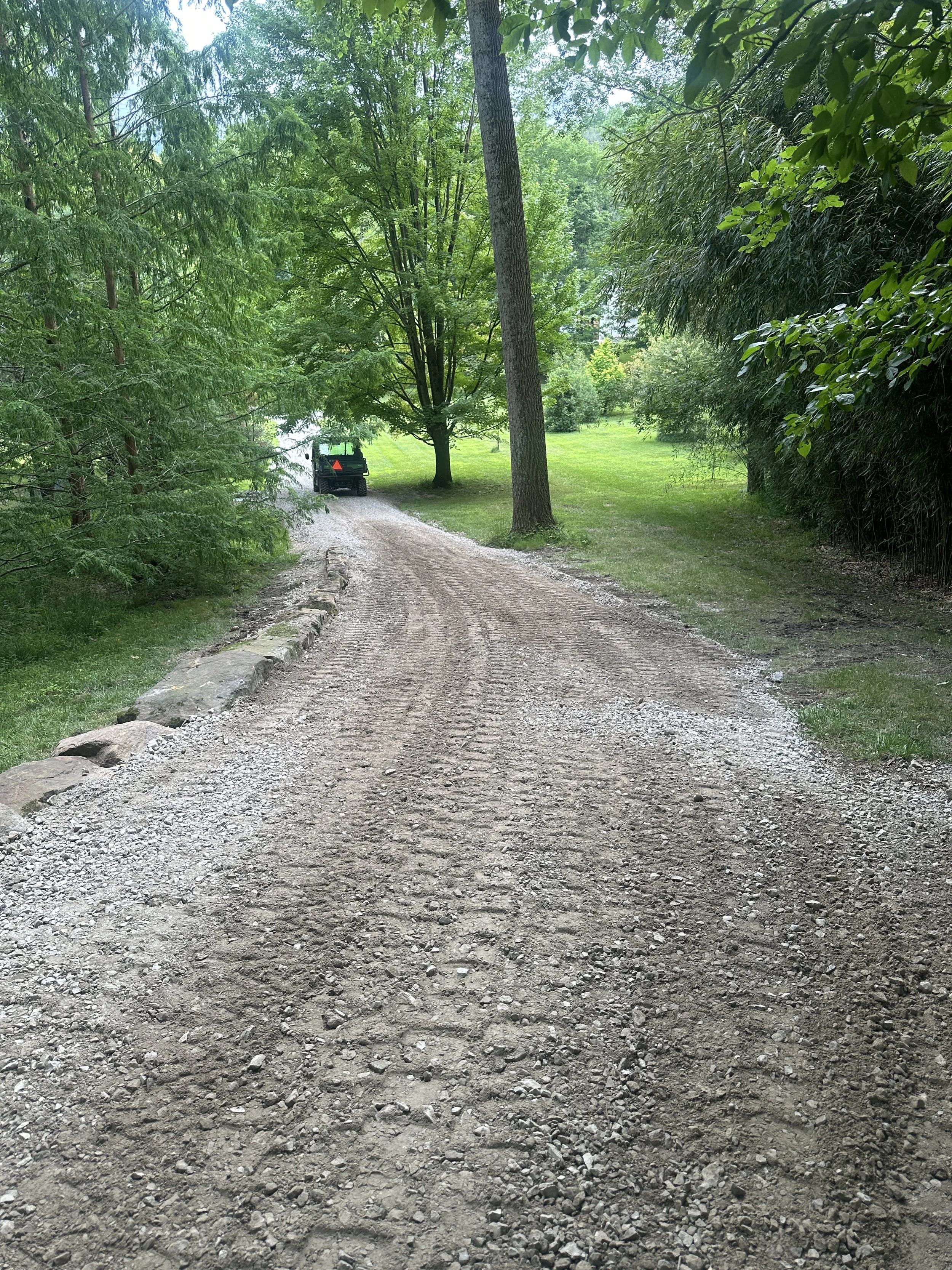 A dirt and gravel road lined with rocks on the left side, surrounded by tall green trees and bushes, with a vehicle visible in the distance on the road.