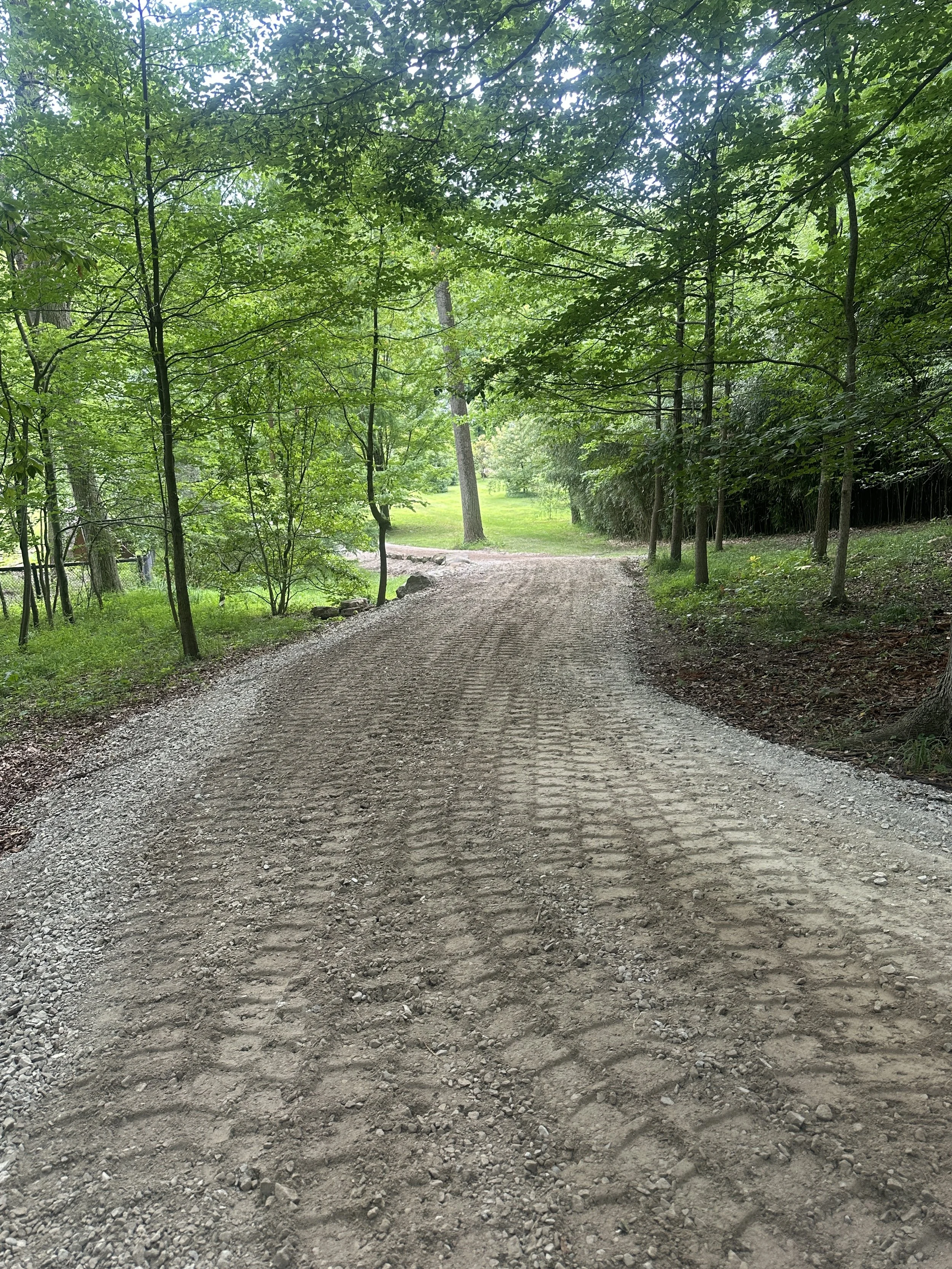 A dirt and gravel pathway surrounded by lush green trees and grass, winding through a forested area.