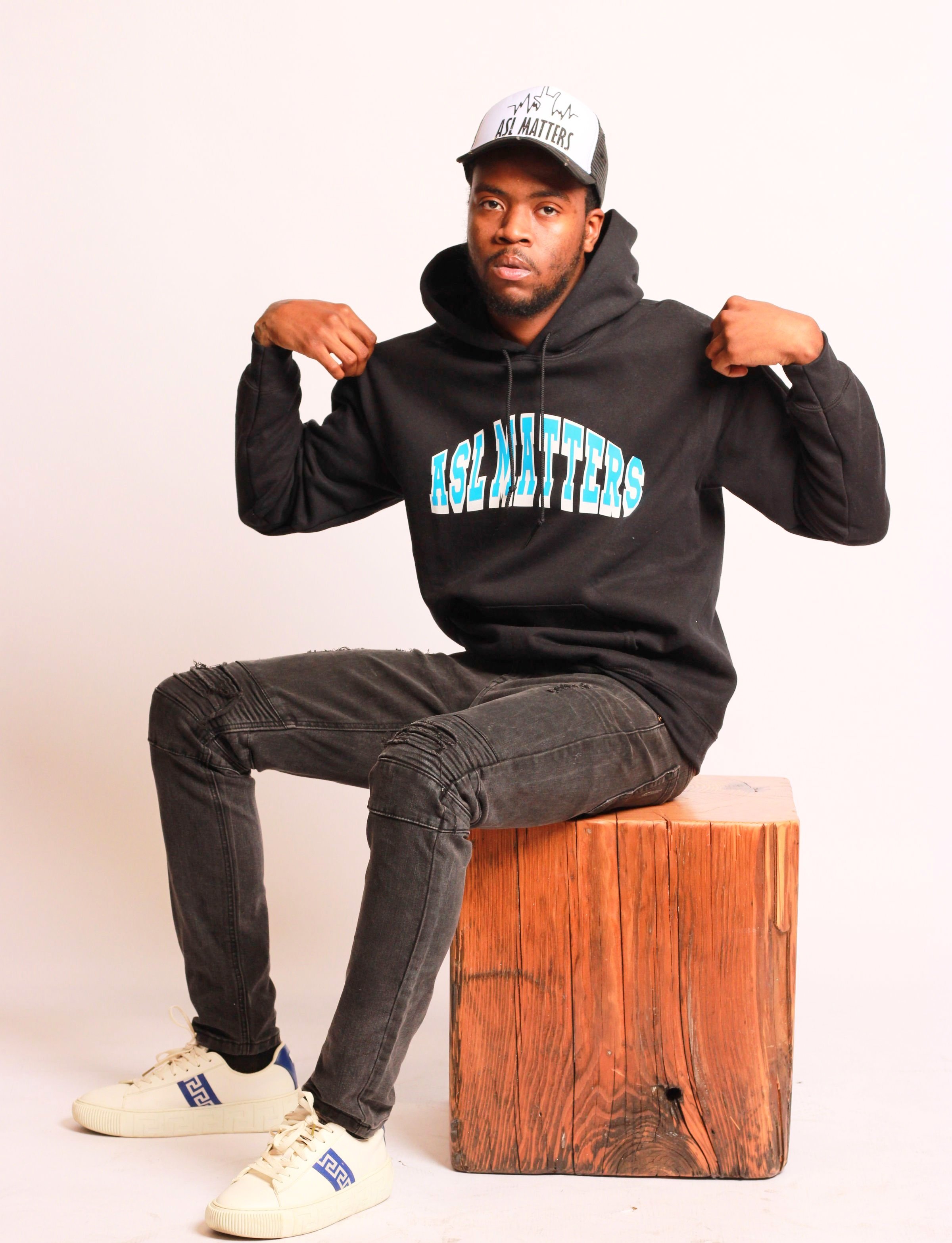 Man wearing a black hoodie with 'ASL MATTERS' text, sitting on a wooden block, wearing a white cap and white sneakers, against a plain background.