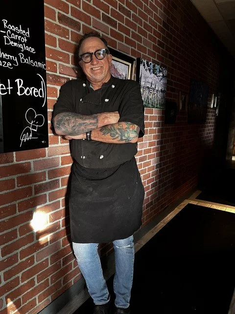 Chef Martin Corso in a black chef's uniform and apron with colorful tattoos on his left arm, standing against a brick wall inside the 'Ronis West restaurant.