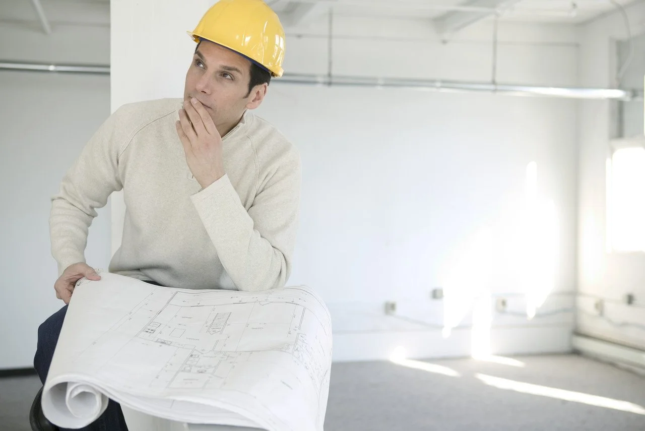 Man wearing a yellow safety helmet holding blueprints and thinking in an unfinished room.