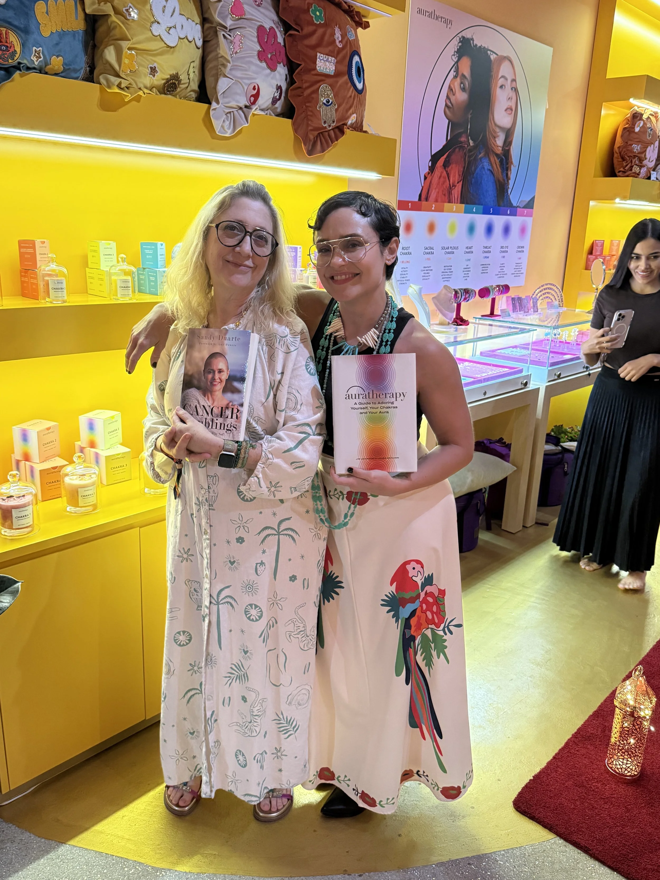 Authors Sandy Duarte and Laura McCann at a Miami event holding their books Cancer Ramblings and Auratherapy, sharing a message of cancer survival, healing, and wellness