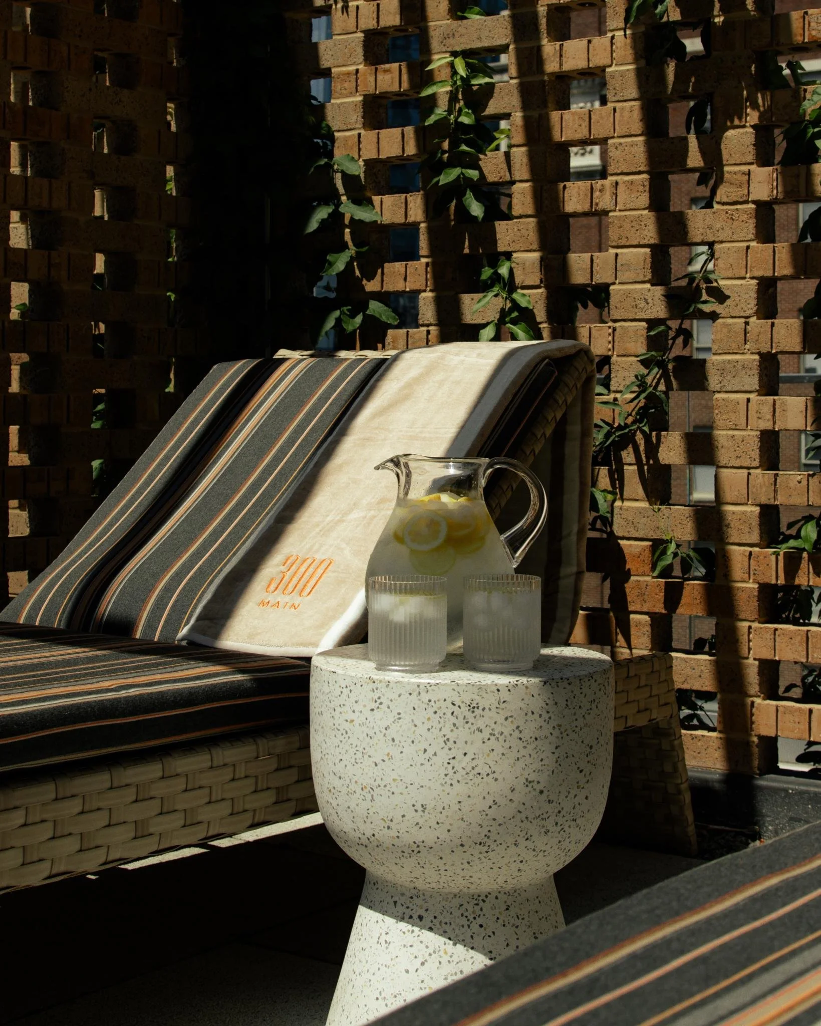 Outdoor lounge seating with striped cushions and a pitcher of lemon water on a small table, set against a textured brick privacy wall at 300 Main.