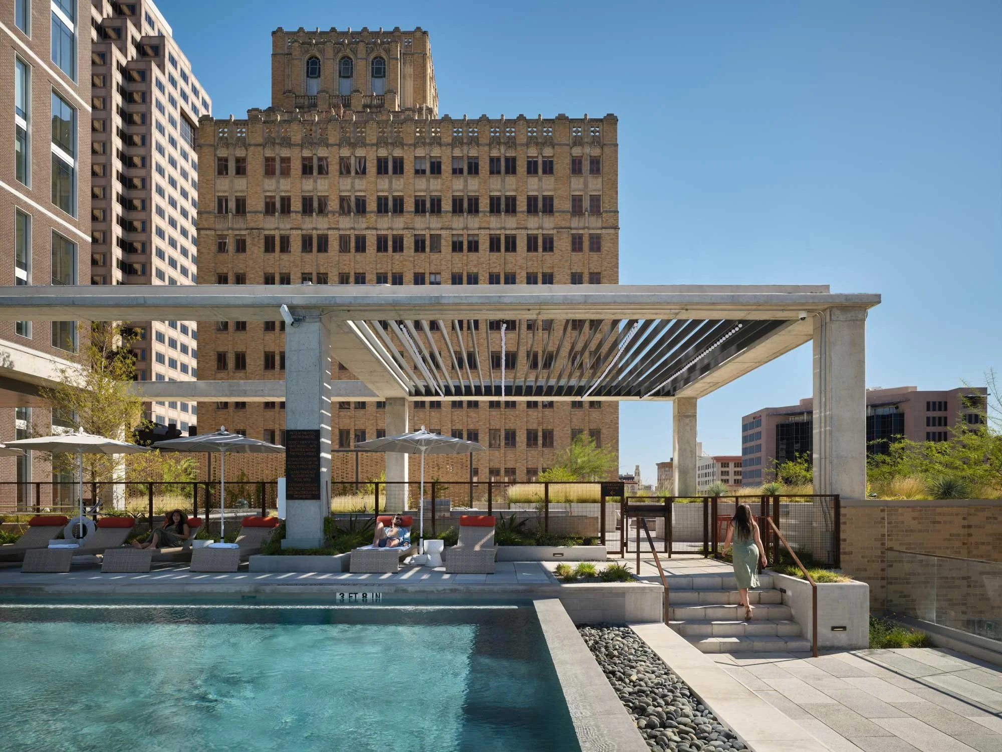Rooftop pool area with lounge chairs, shaded pergola, and city skyline views behind a vintage high-rise building.