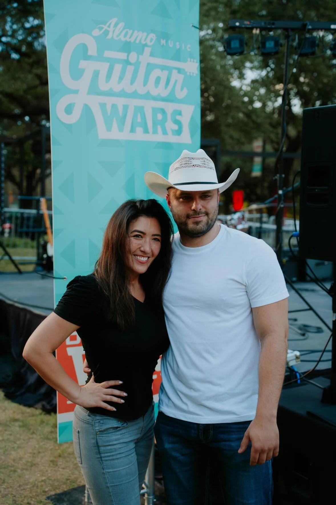 Two people posing and smiling in front of a Guitar Wars event sign at an outdoor music festival.