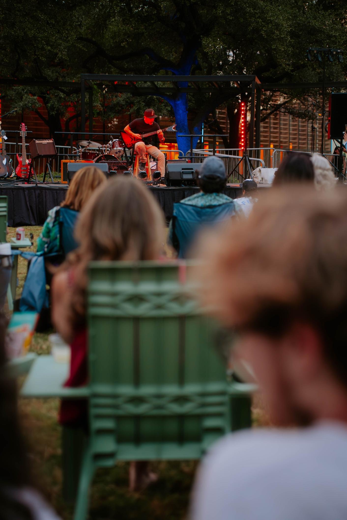 Outdoor concert scene with a guitarist performing on stage and audience members seated in lawn chairs.