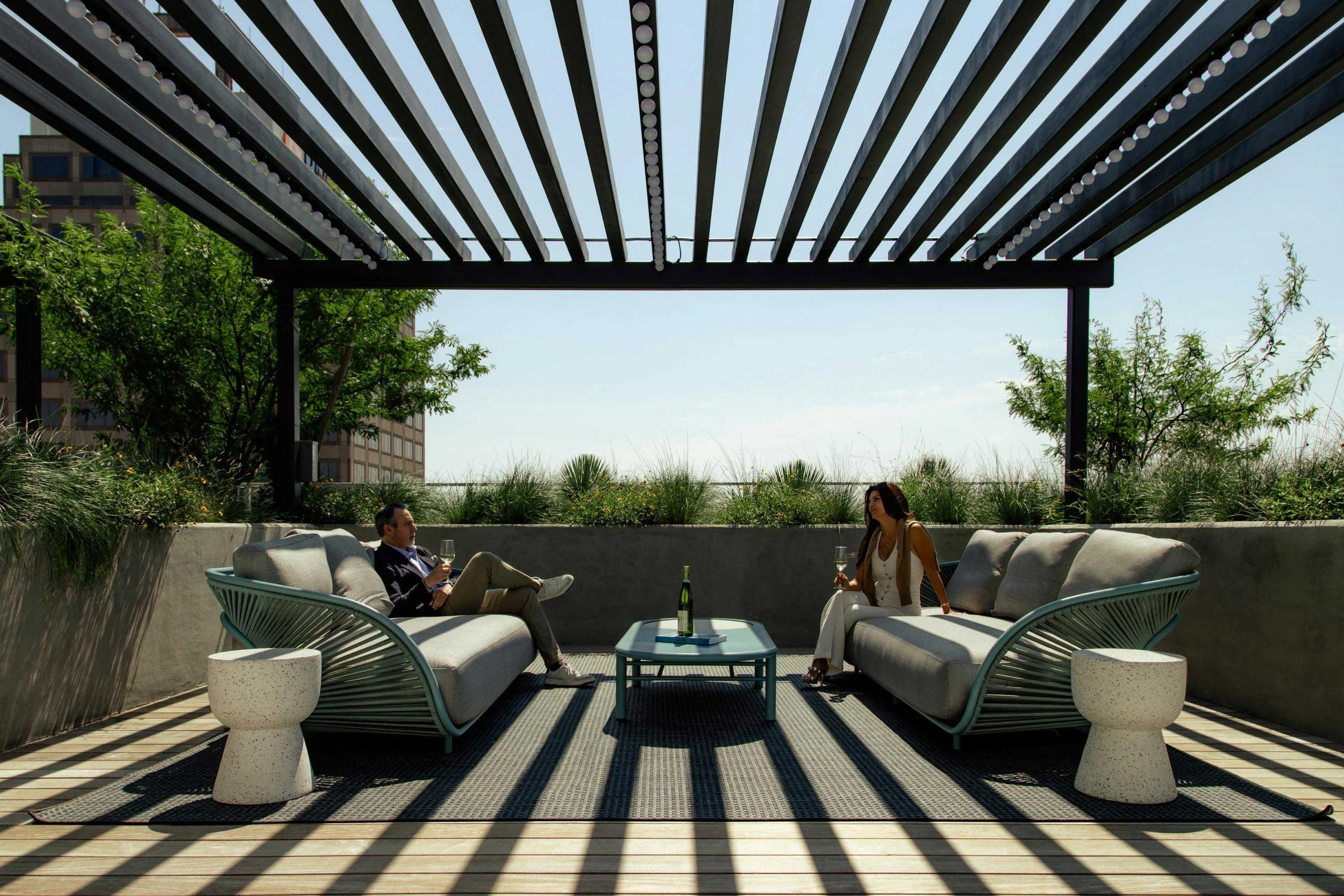 Two people sitting on outdoor sofas under a pergola on a rooftop lounge, enjoying drinks with a low table between them and greenery behind.