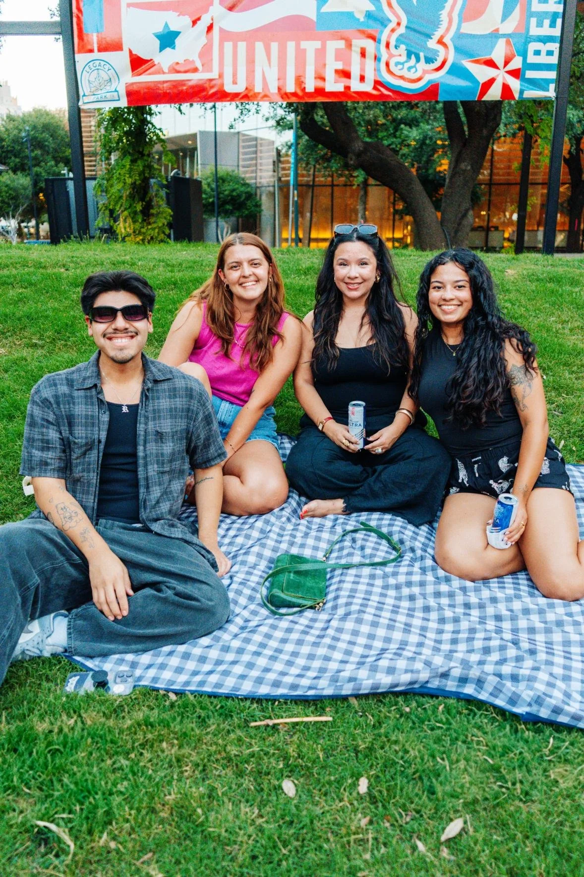 Group of four friends sitting on a picnic blanket on the grass under a United banner at an outdoor event.