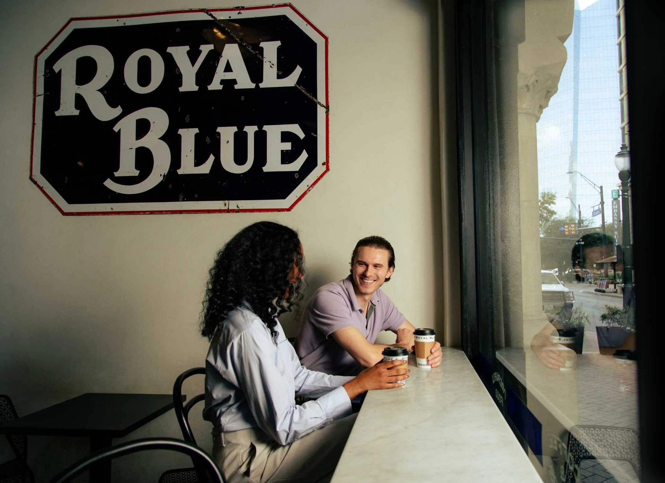 Two people sitting in a cafe near a window drinking coffee with a "Royal Blue" sign on the wall.