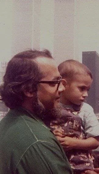 A  bearded man holds a child with the twin towers of the World Trade Center visible behind them