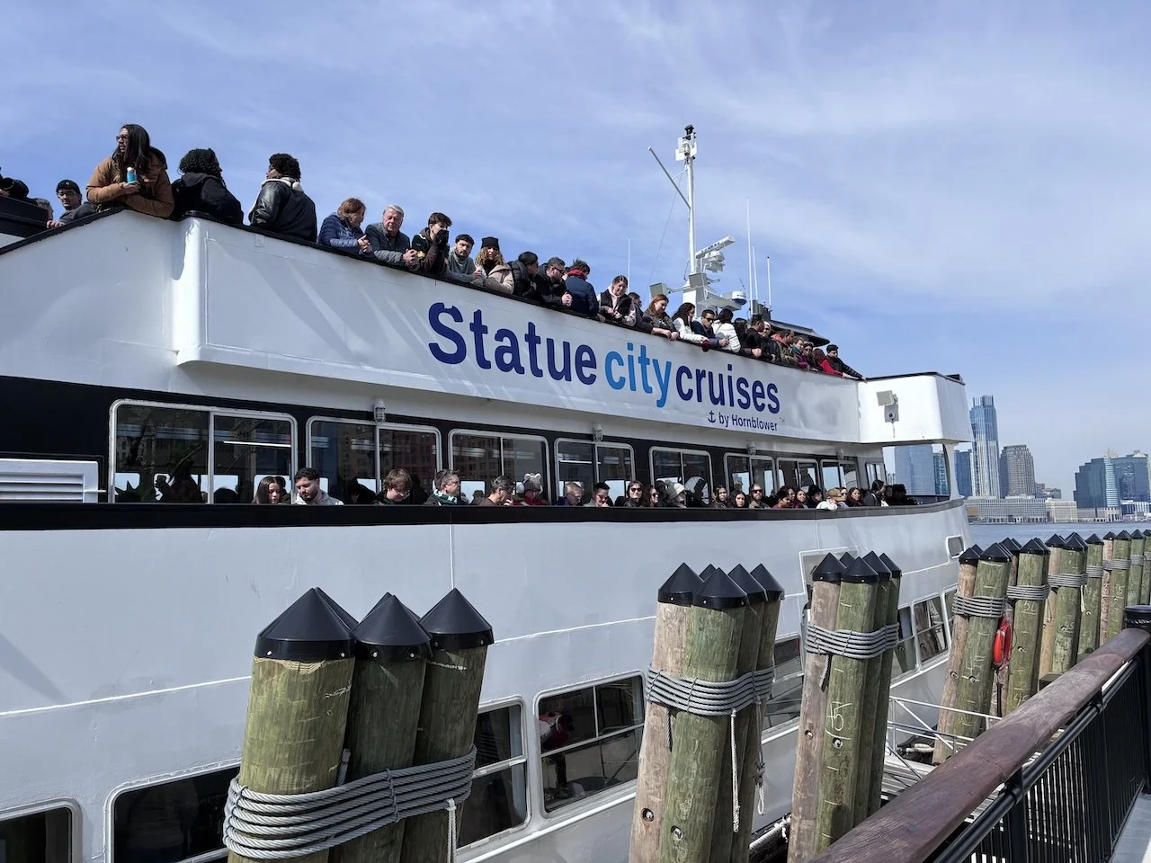 White ferry boat packed with passengers. Side of boat is labeled Statue City Cruises by Hornblower
