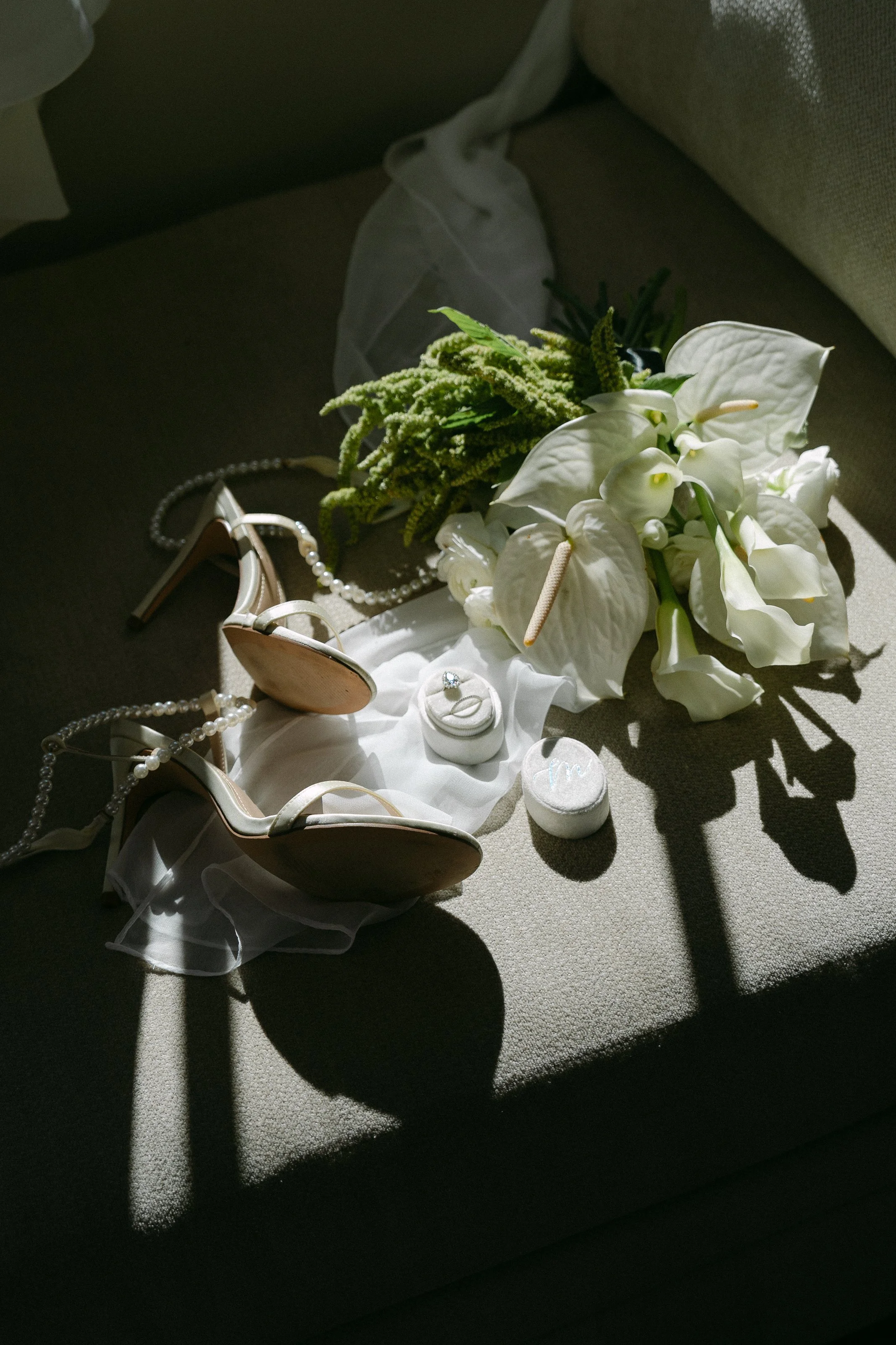Wedding accessories arranged on a couch, including white high heels, a pearl necklace, a bouquet of white anthuriums and greenery, a diamond ring, and a small container of pills, with sunlight creating shadows.