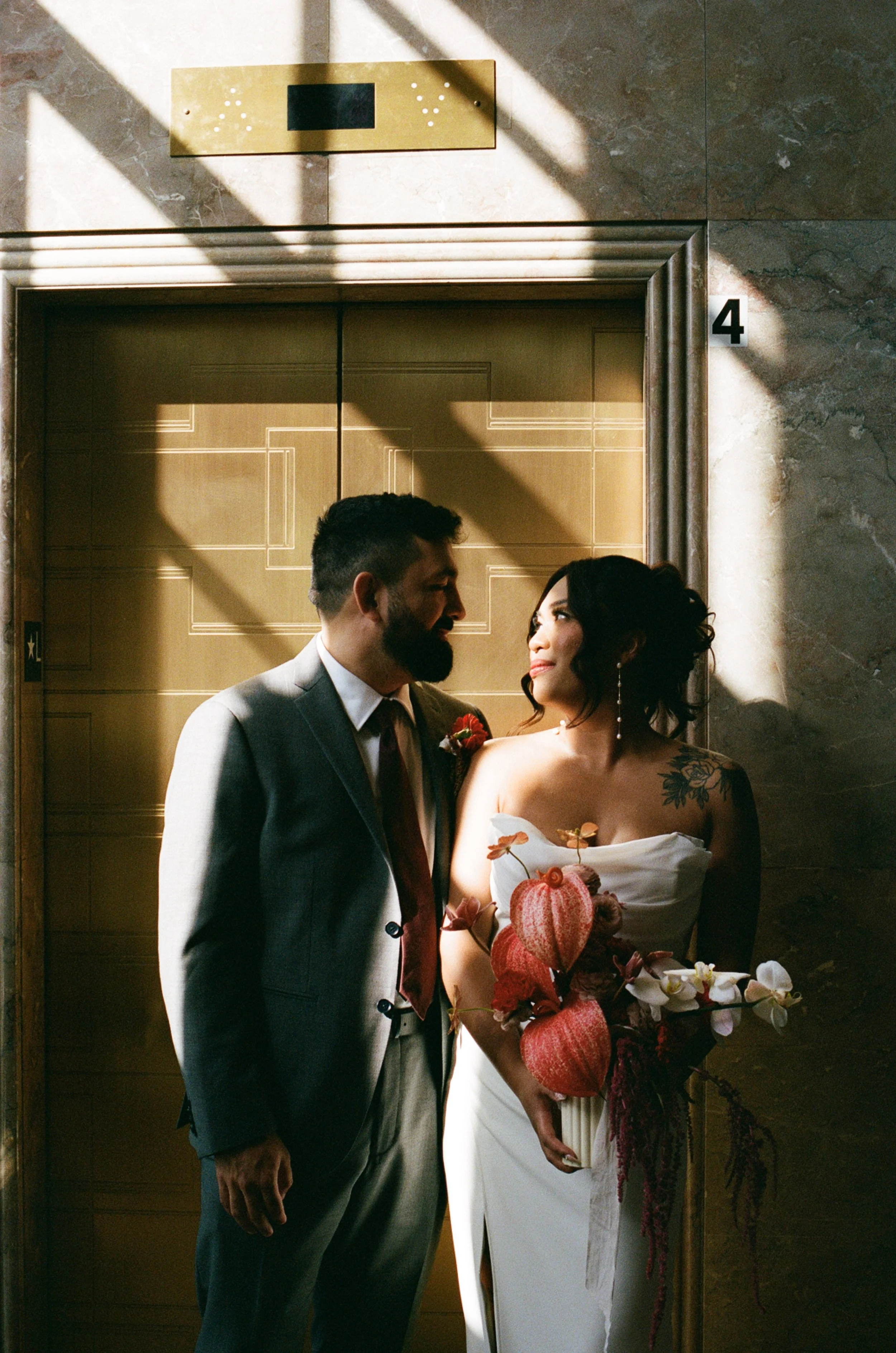 A couple in wedding attire standing close in front of an elevator, with sunlight casting dramatic shadows on the wall.