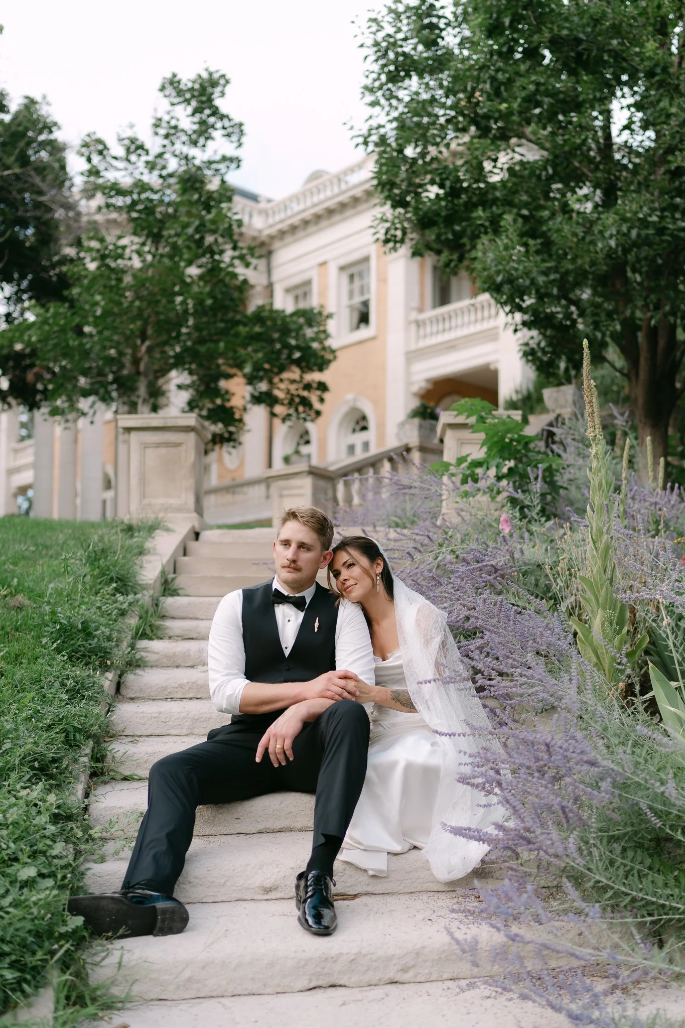 Nika Libres Photo San Diego - A newlywed couple sitting on stone steps outside a large elegant house surrounded by greenery, during daytime.