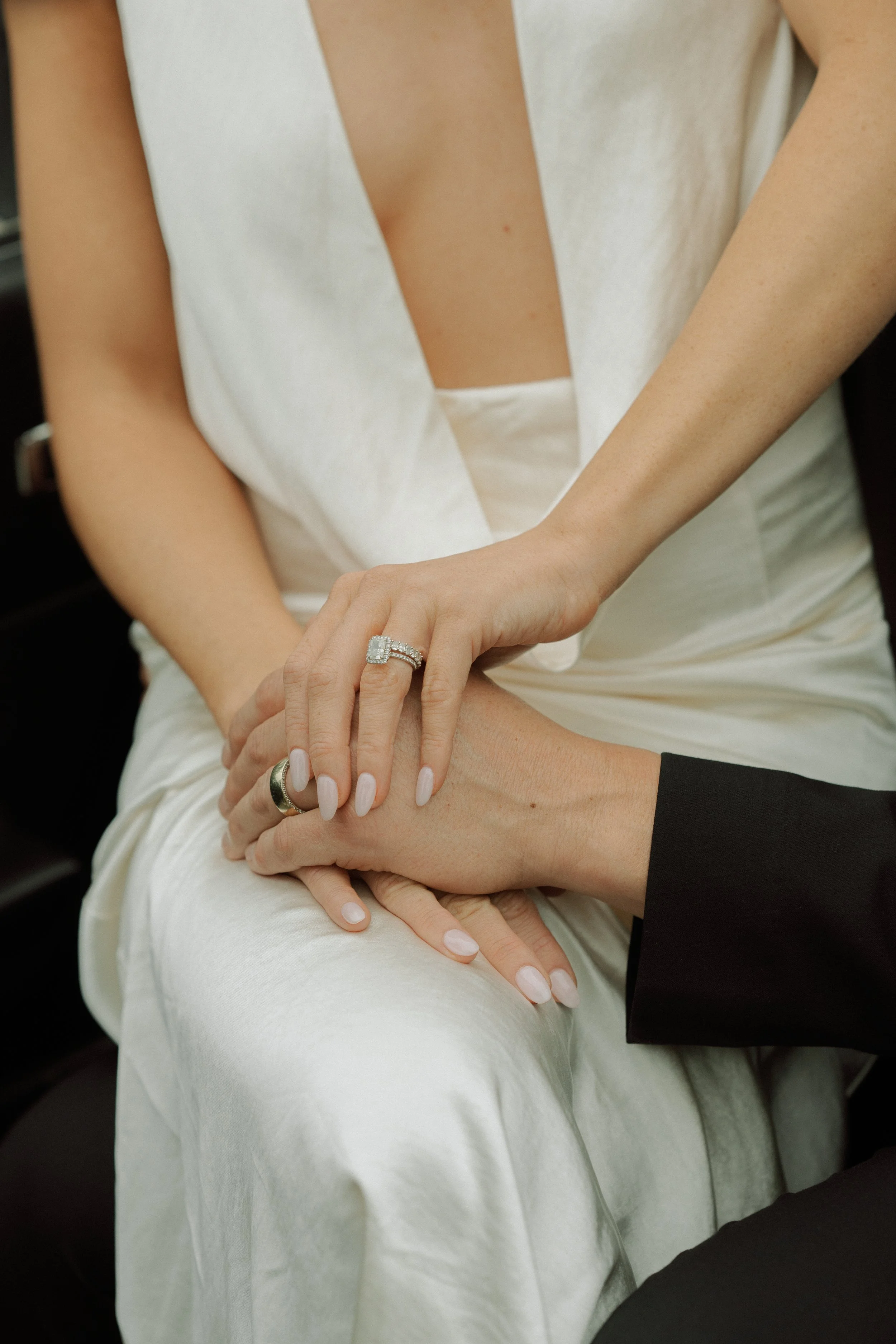 A woman in a white dress and a man in black suit holding hands, showing wedding rings.