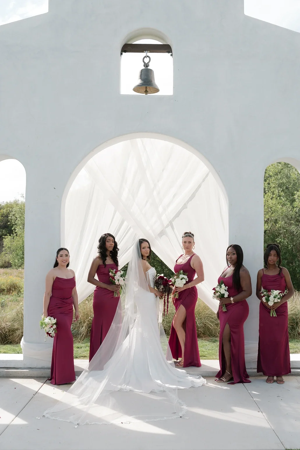 Bride with her bridesmaids in burgundy gowns holding bouquets beneath the draped ceremony arch at Jeune Perché Estate