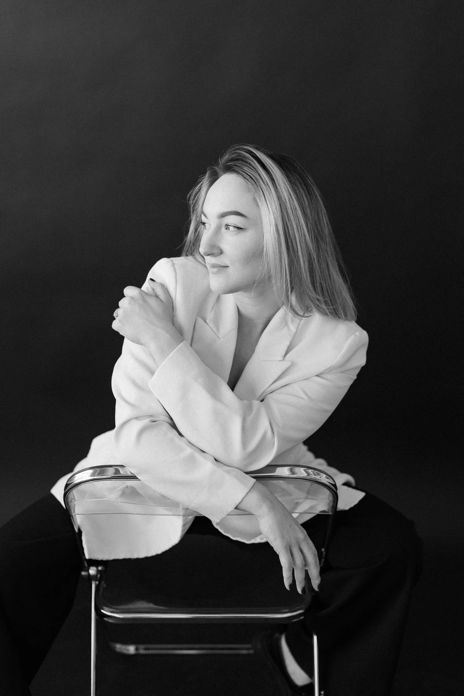 A black and white portrait of a young woman with light hair, sitting on a transparent chair against a dark background. She is wearing a light-colored blazer and dark pants, looking to the side with a calm expression.