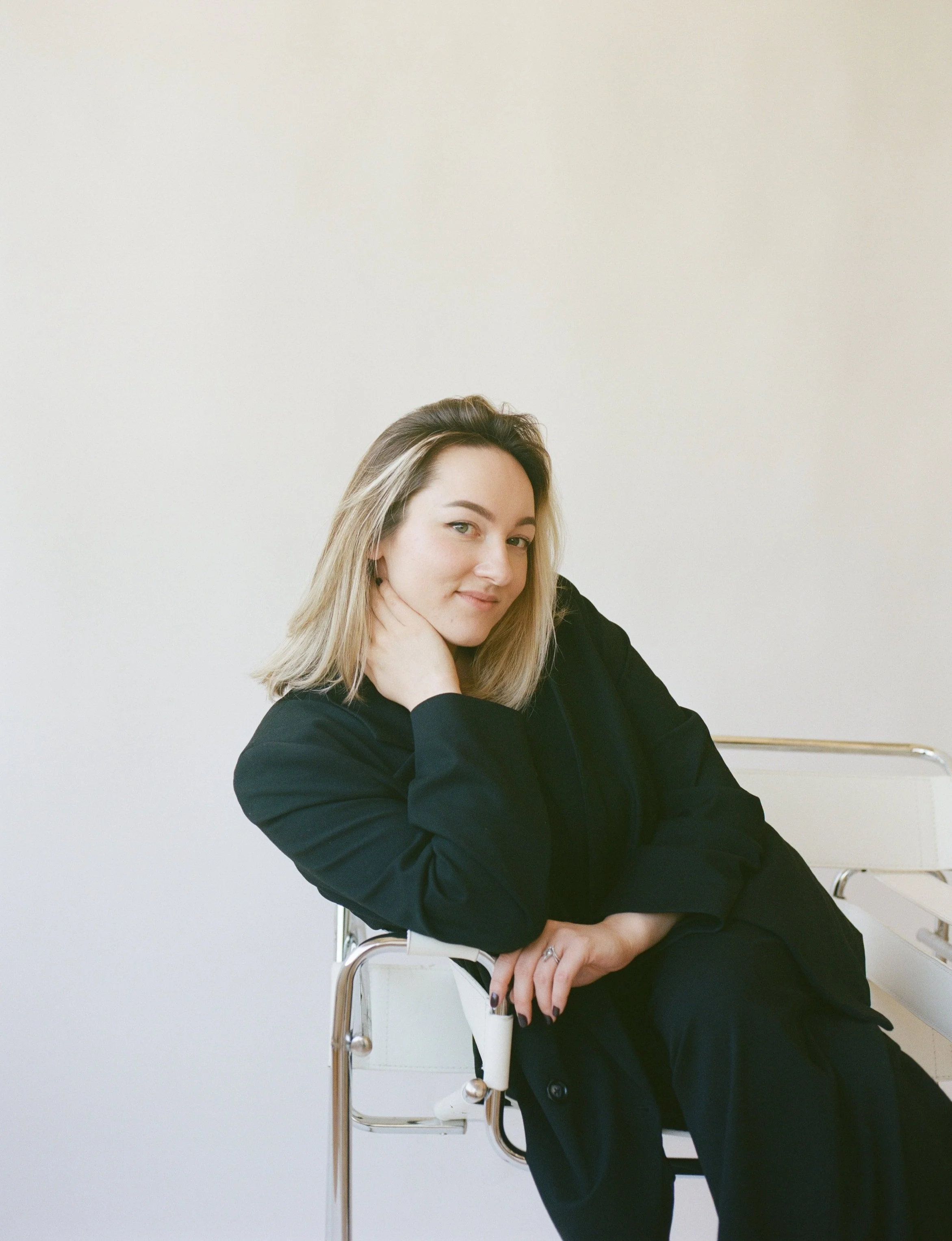 A woman with blonde hair wearing a black suit, sitting on a white chair against a plain light background, looking at the camera with a relaxed expression.