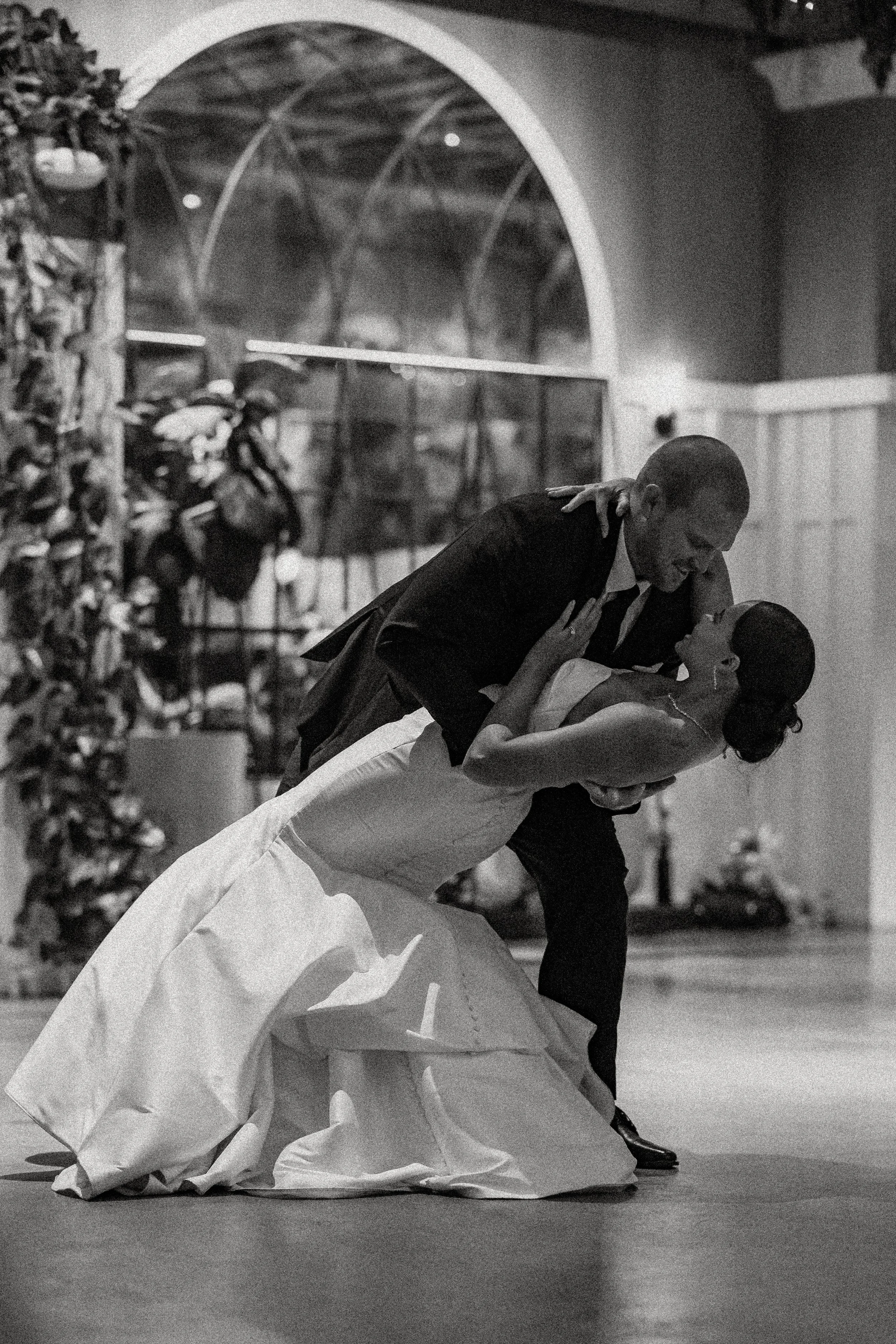 A black-and-white photo of a bride and groom dancing, with the groom dipping the bride, in a decorated indoor setting.