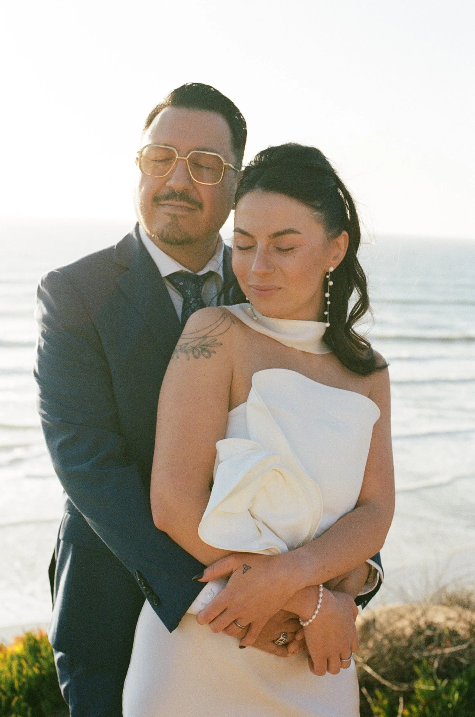 A couple in wedding attire embracing on the beach during sunset, with the ocean in the background.