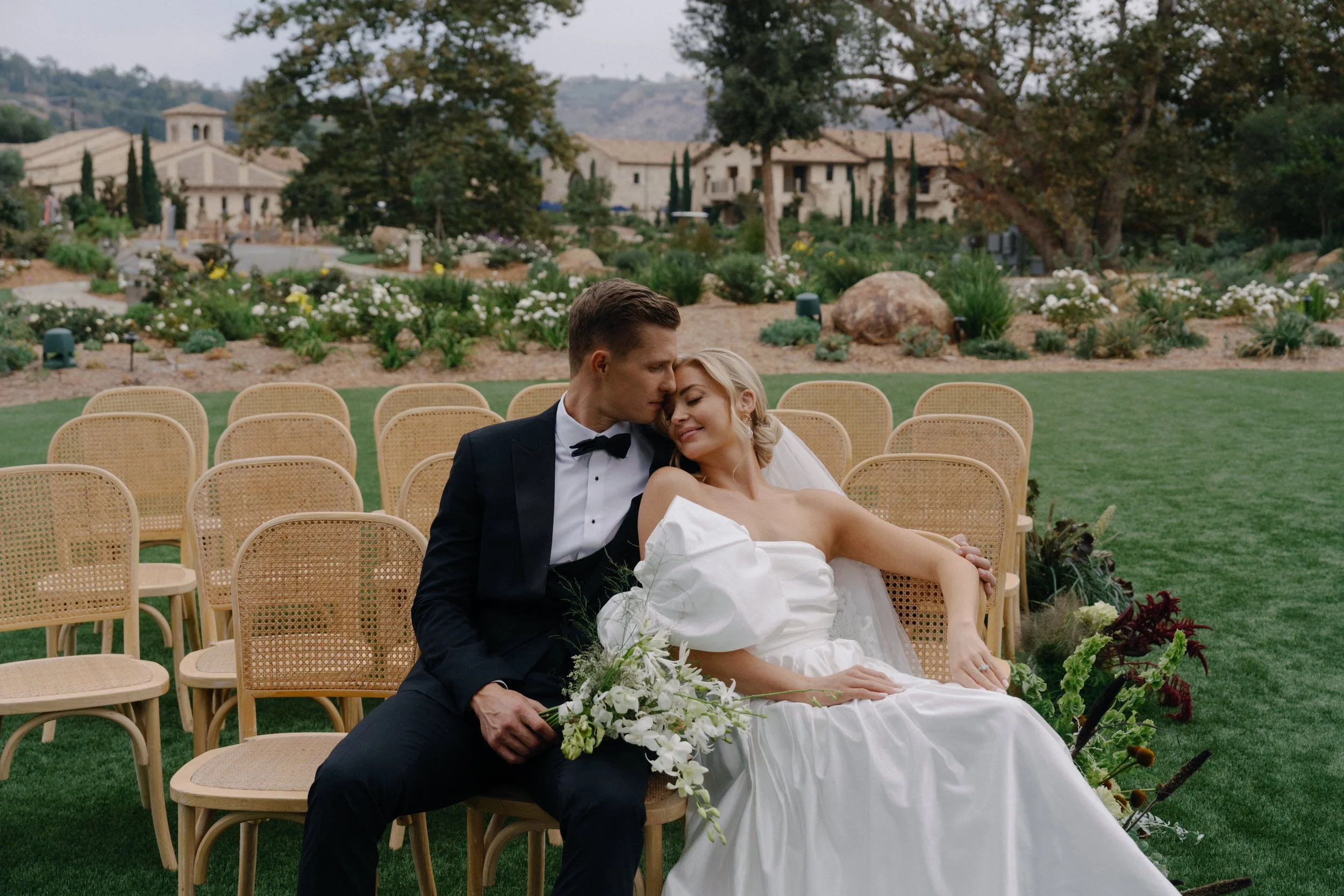 A bride and groom sitting closely together on wooden chairs outdoors during a wedding ceremony, with chairs arranged in rows behind them, a well-manicured lawn, and a lush garden with trees and flowers in the background.