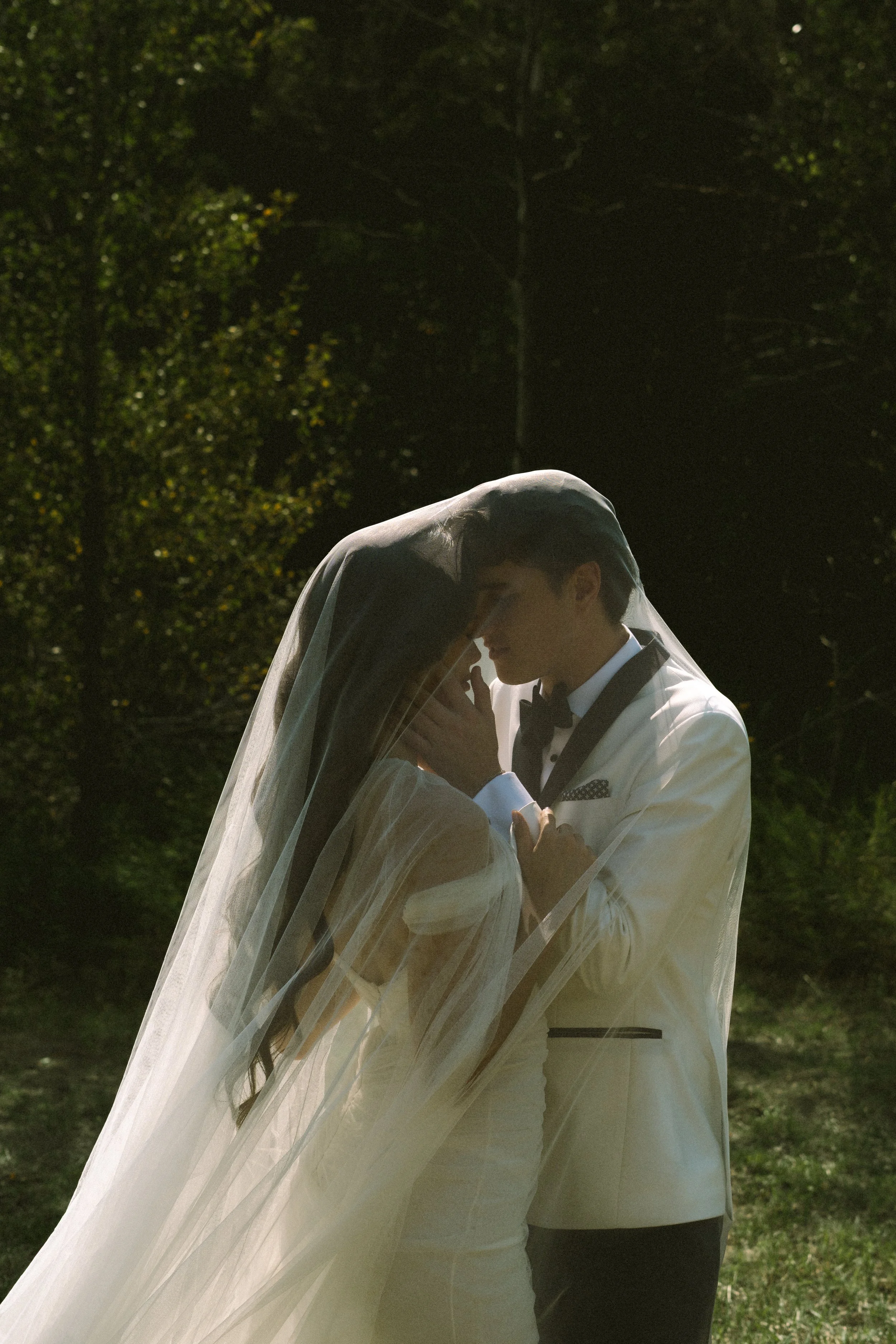 A bride and groom sharing an intimate moment outdoors, with the bride wearing a veil and a white dress, and the groom in a white tuxedo with a black bow tie, surrounded by greenery.