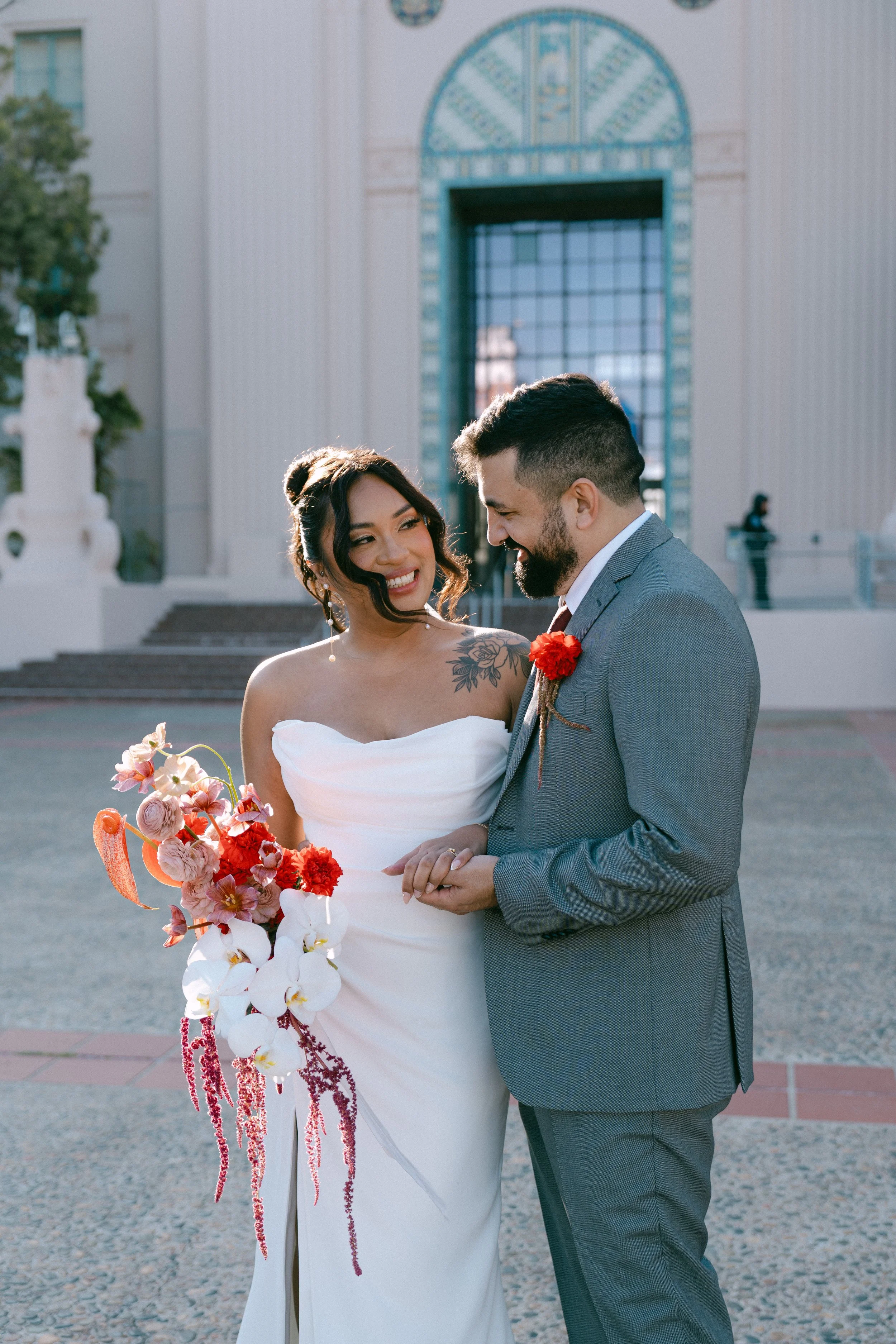 A bride and groom on their wedding day embrace and smile at each other outdoors, with a building in the background.