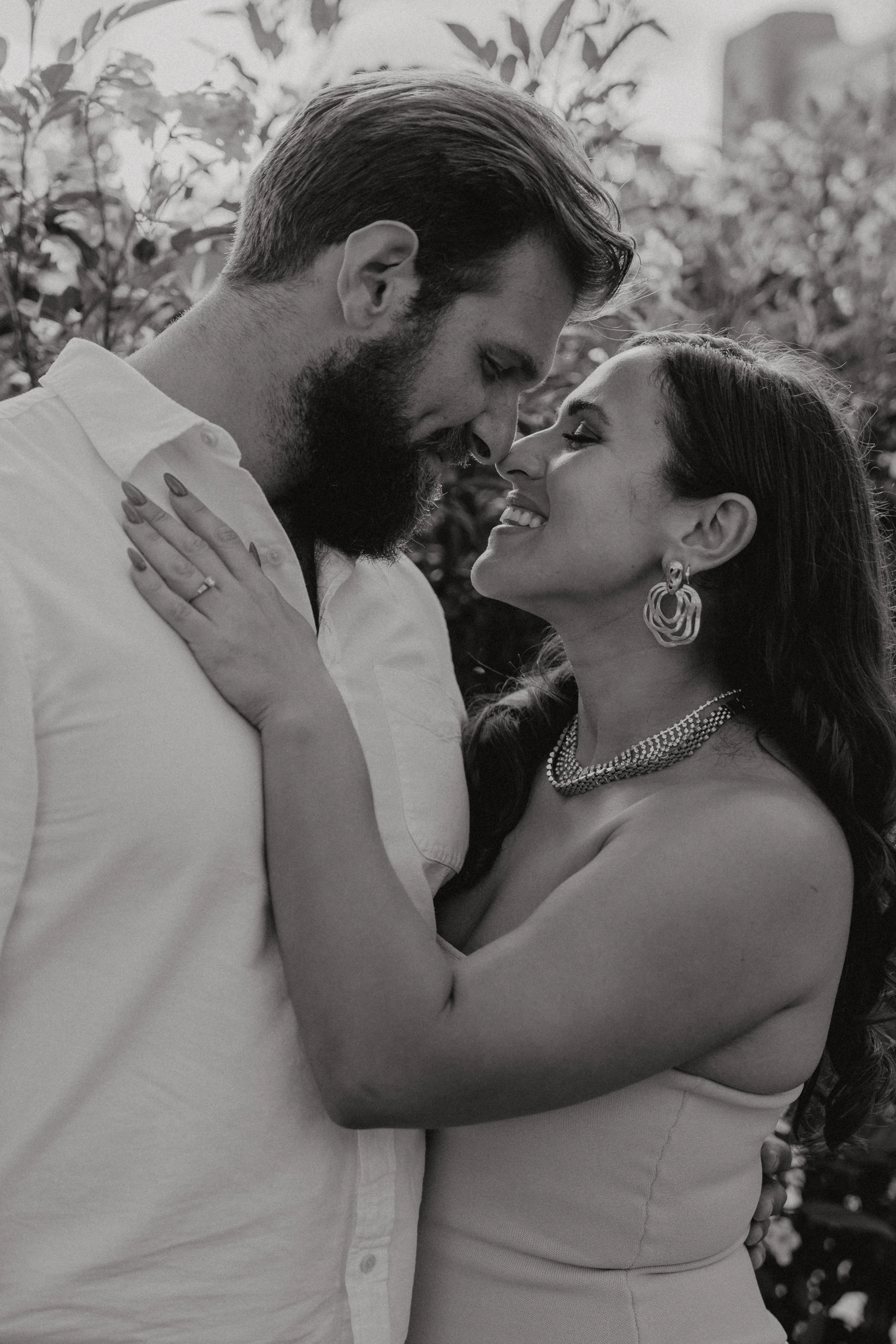 A black and white photo of a loving couple standing close together outdoors, with their foreheads touching and smiling at each other, surrounded by trees and foliage.