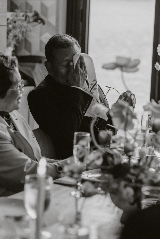 An elderly man in a suit is wiping his eyes with a napkin at a dining table, possibly emotional. Two women are sitting beside him, and the table is set with glasses, plates, and floral decorations. The scene appears to be in a well-lit room near a window.