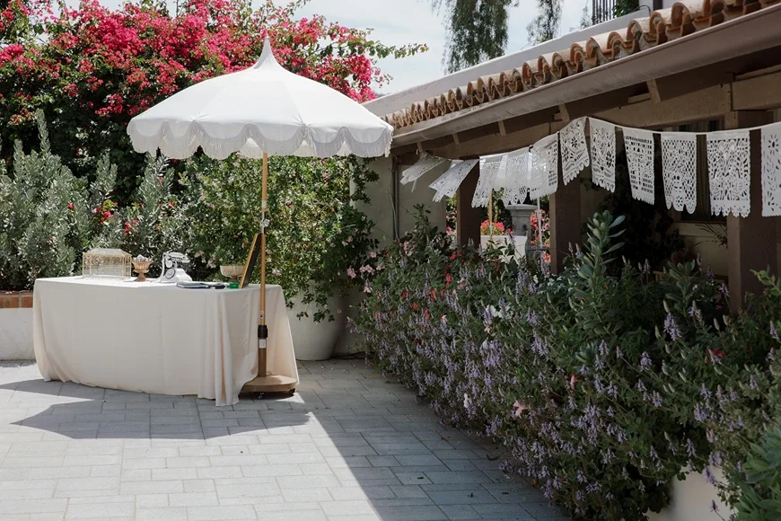 Outdoor welcome area at Jeune Perché Estate with a scalloped white umbrella, draped table, and papel picado banners among blooming bougainvillea