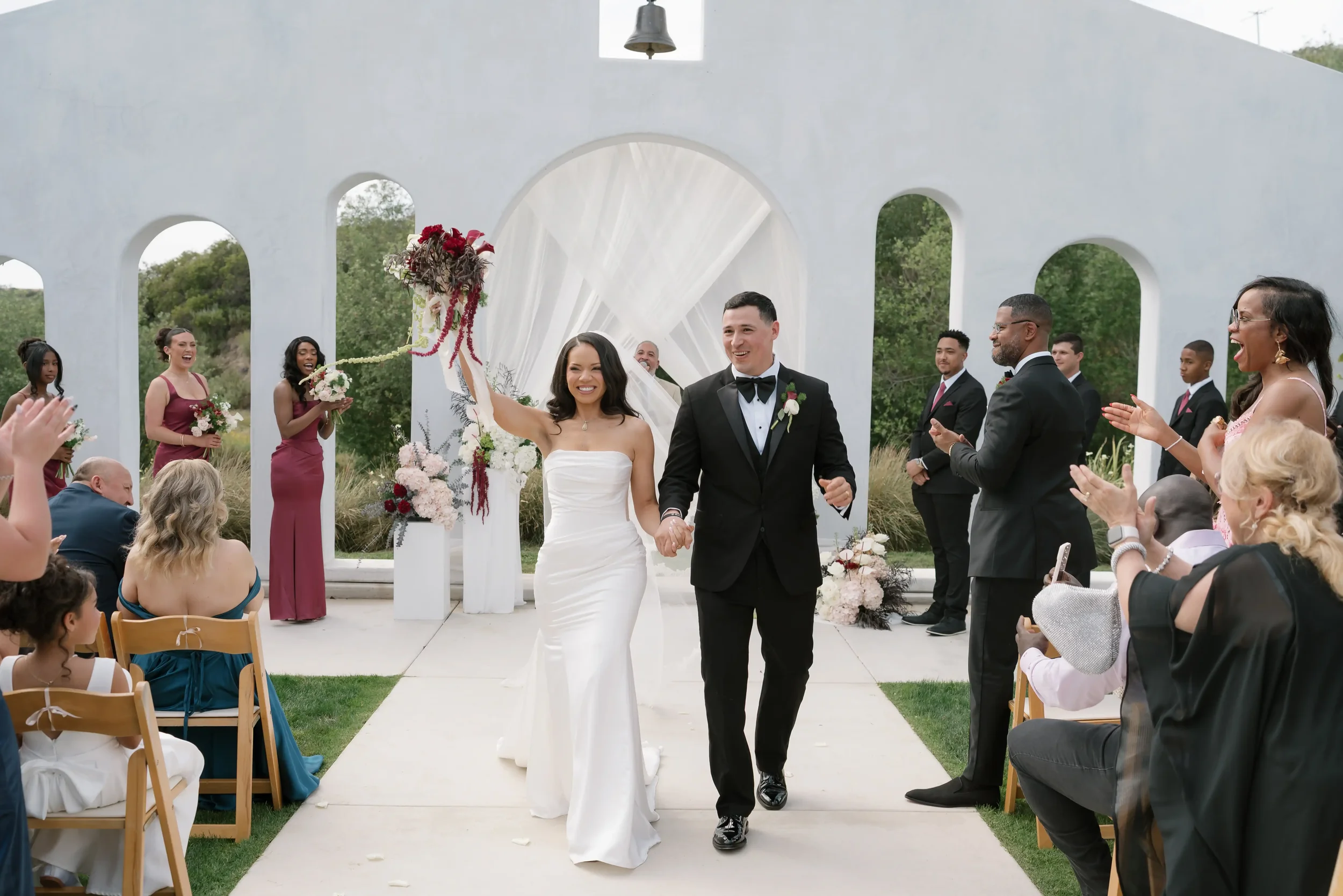 Bride and groom walking back up the aisle as guests cheer at their Jeune Perché Estate wedding ceremony, in front of a draped arch
