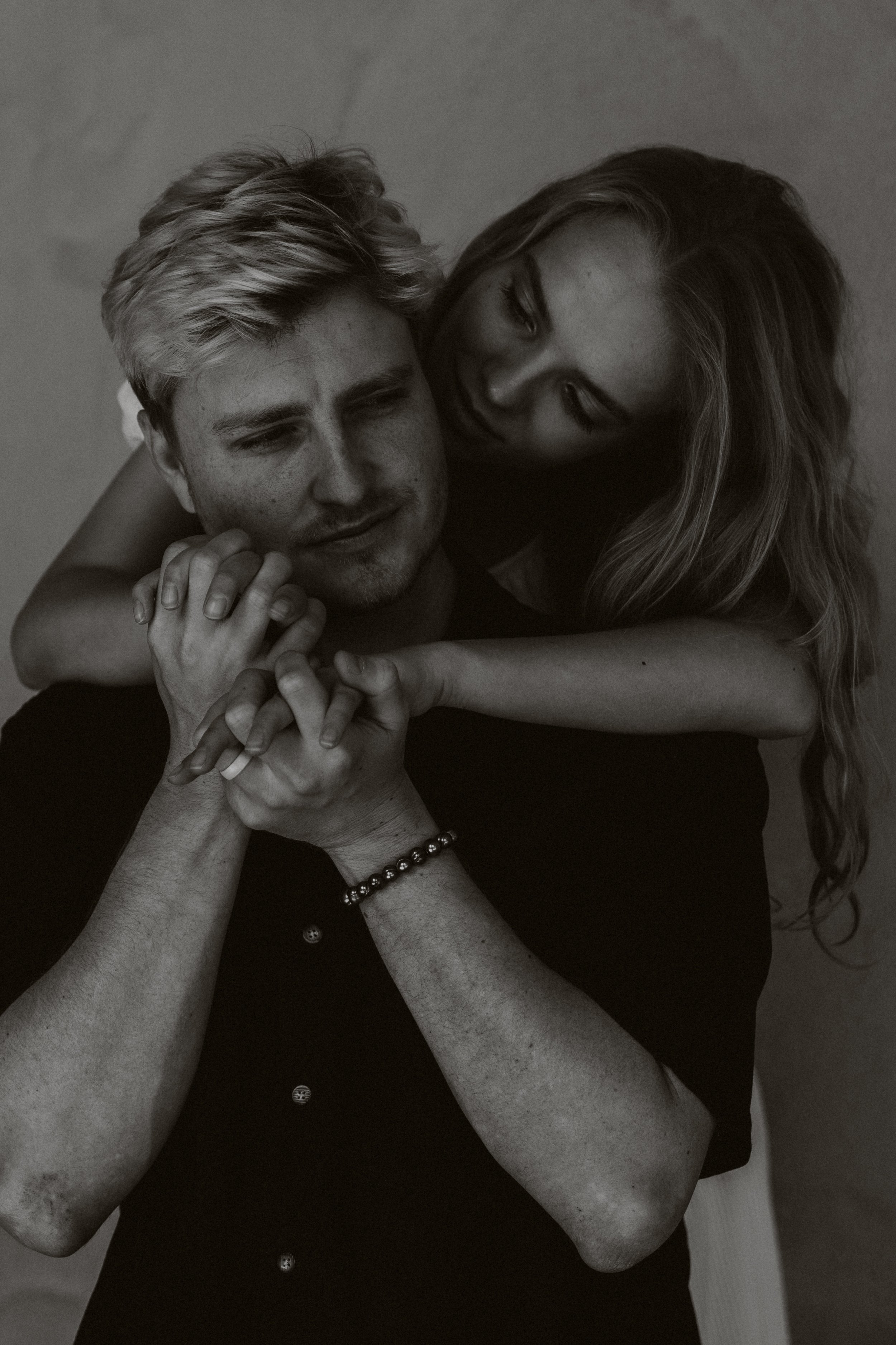 Nika Libres Photo San Diego - 
A black and white photo of a young man and woman in an embrace, with the woman hugging the man from behind. They are holding hands and appear to be close and intimate.