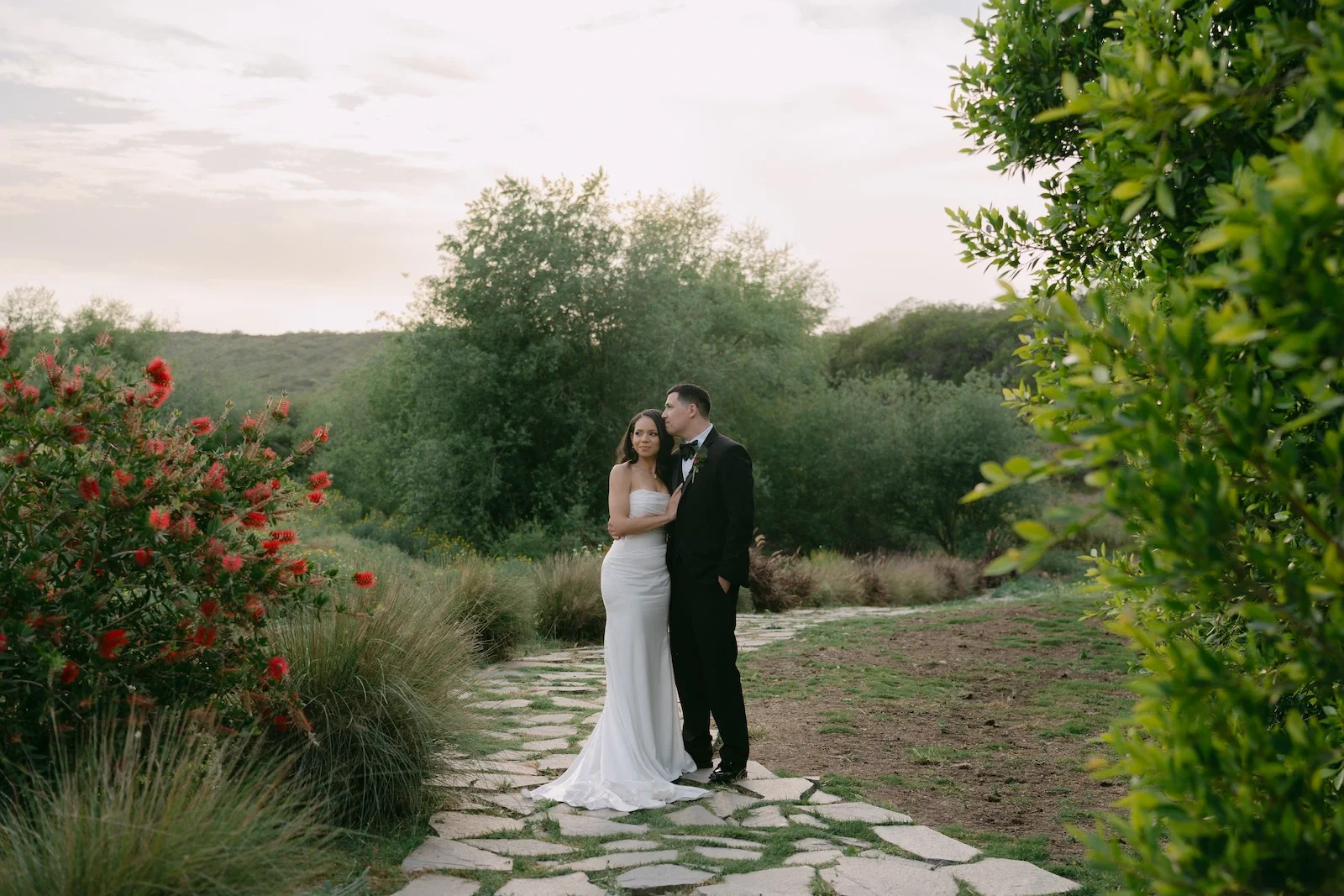 Bride and groom in a romantic embrace on a stone path surrounded by greenery and red blooms during golden-hour sunset portraits at Jeune Perché Estate