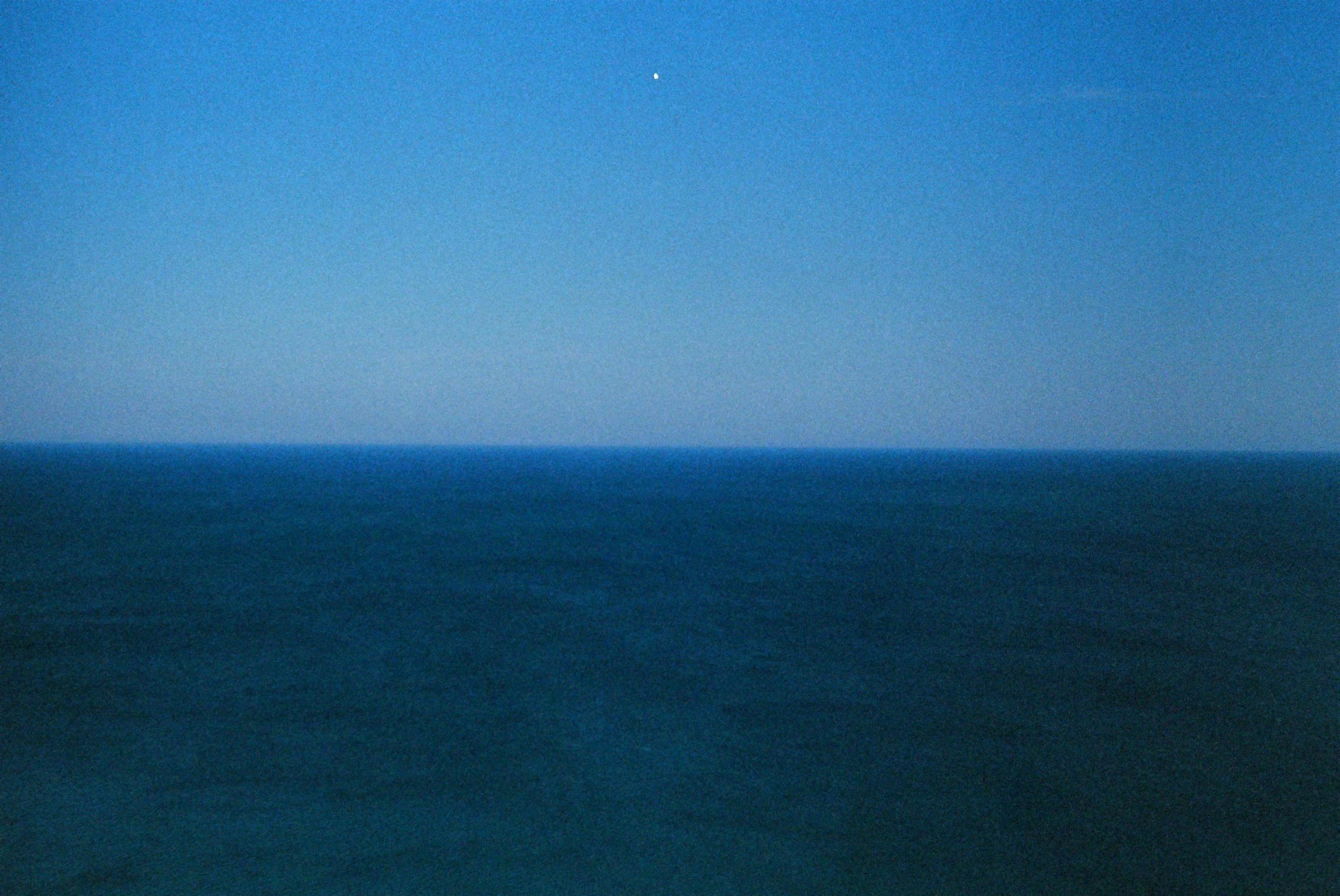 A vast ocean under a clear blue sky with a tiny white dot, possibly an aircraft or bird, visible near the top center of the image.