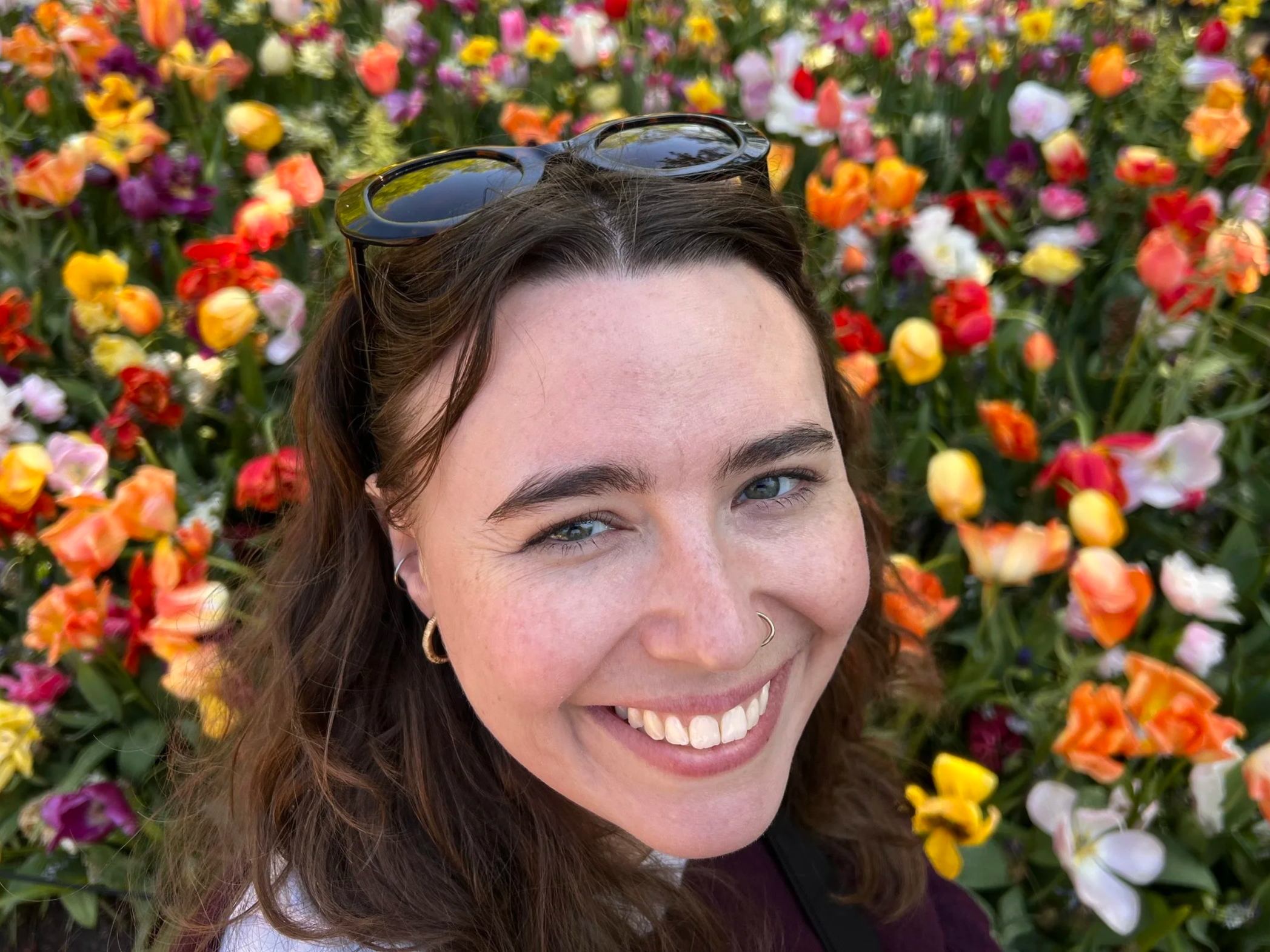 Close-up photo of a smiling woman with brown hair and glasses on her head, surrounded by a colorful array of flowers.