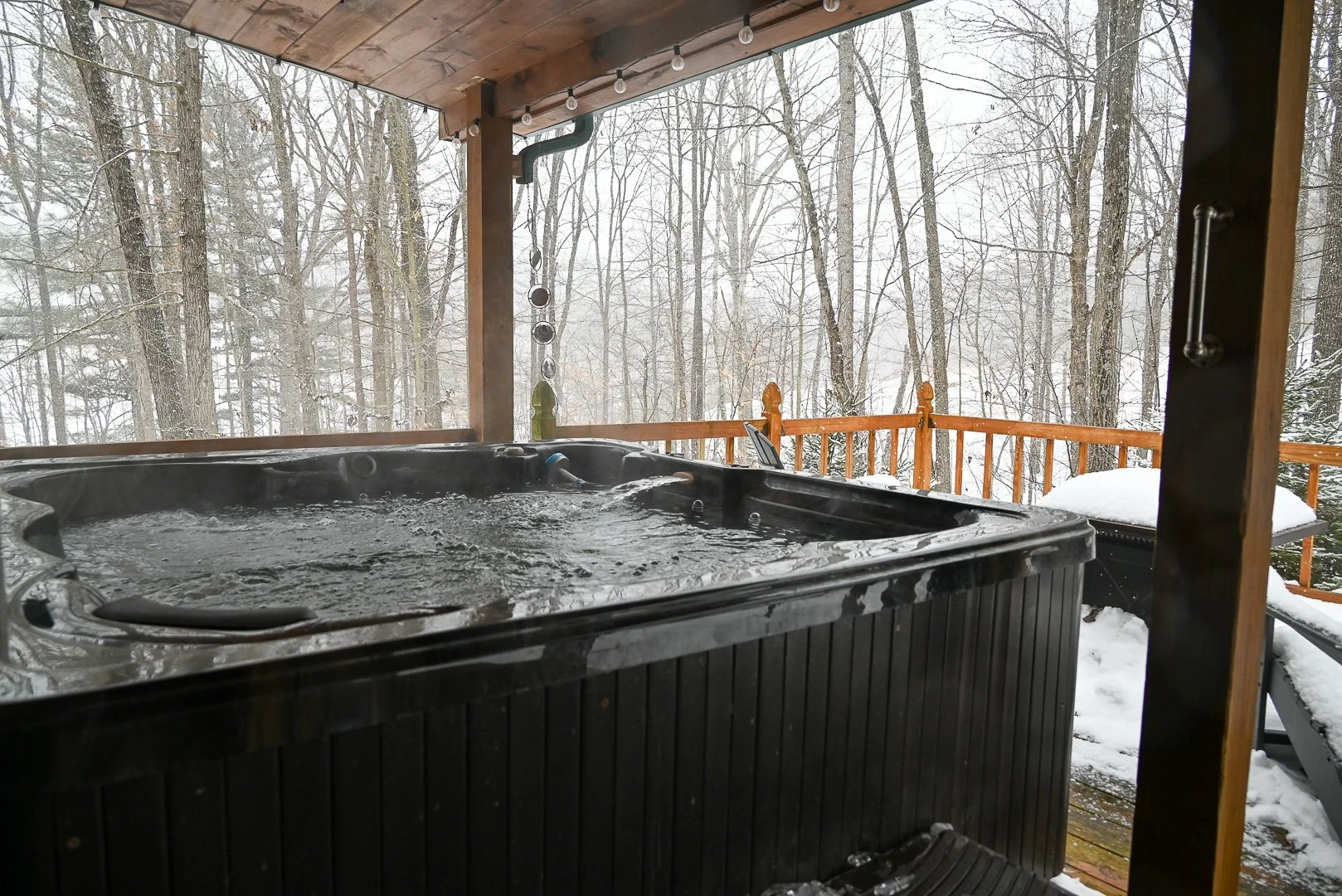 A steaming hot tub on a covered cabin deck overlooking snow‑covered woods in Hocking Hills, creating a peaceful winter nature escape.