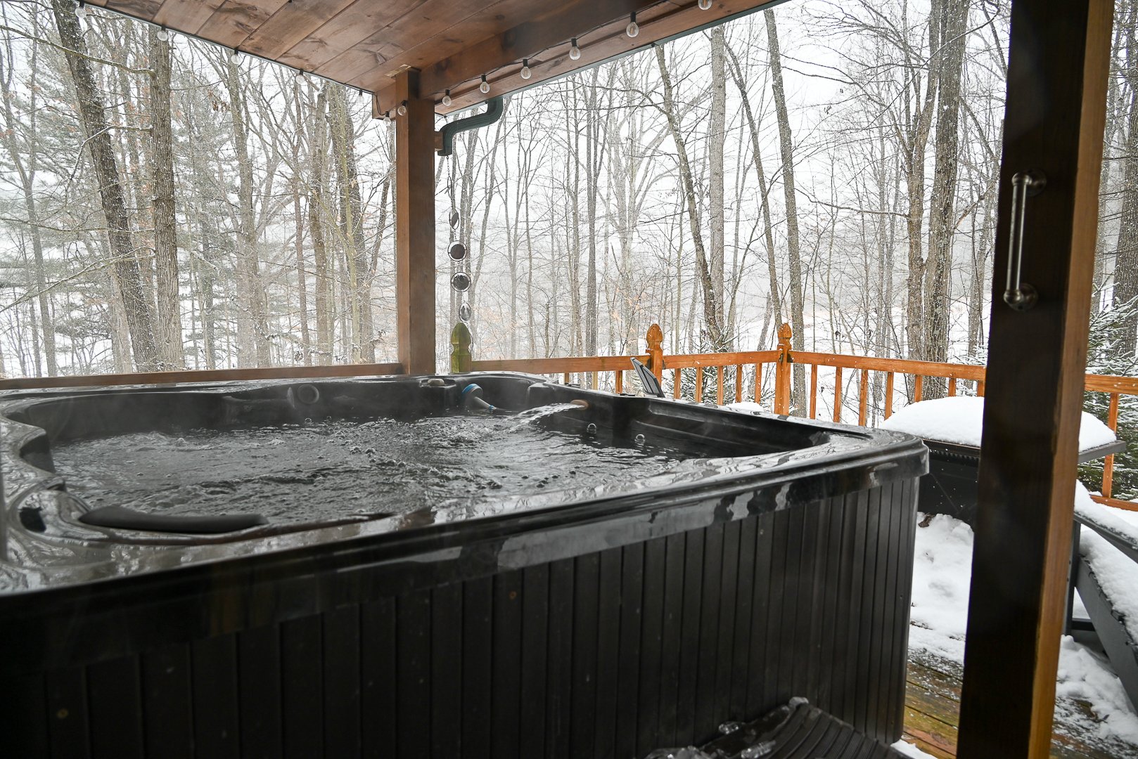 A steaming hot tub on a covered cabin deck overlooking snow‑covered woods in Hocking Hills, creating a peaceful winter nature escape.