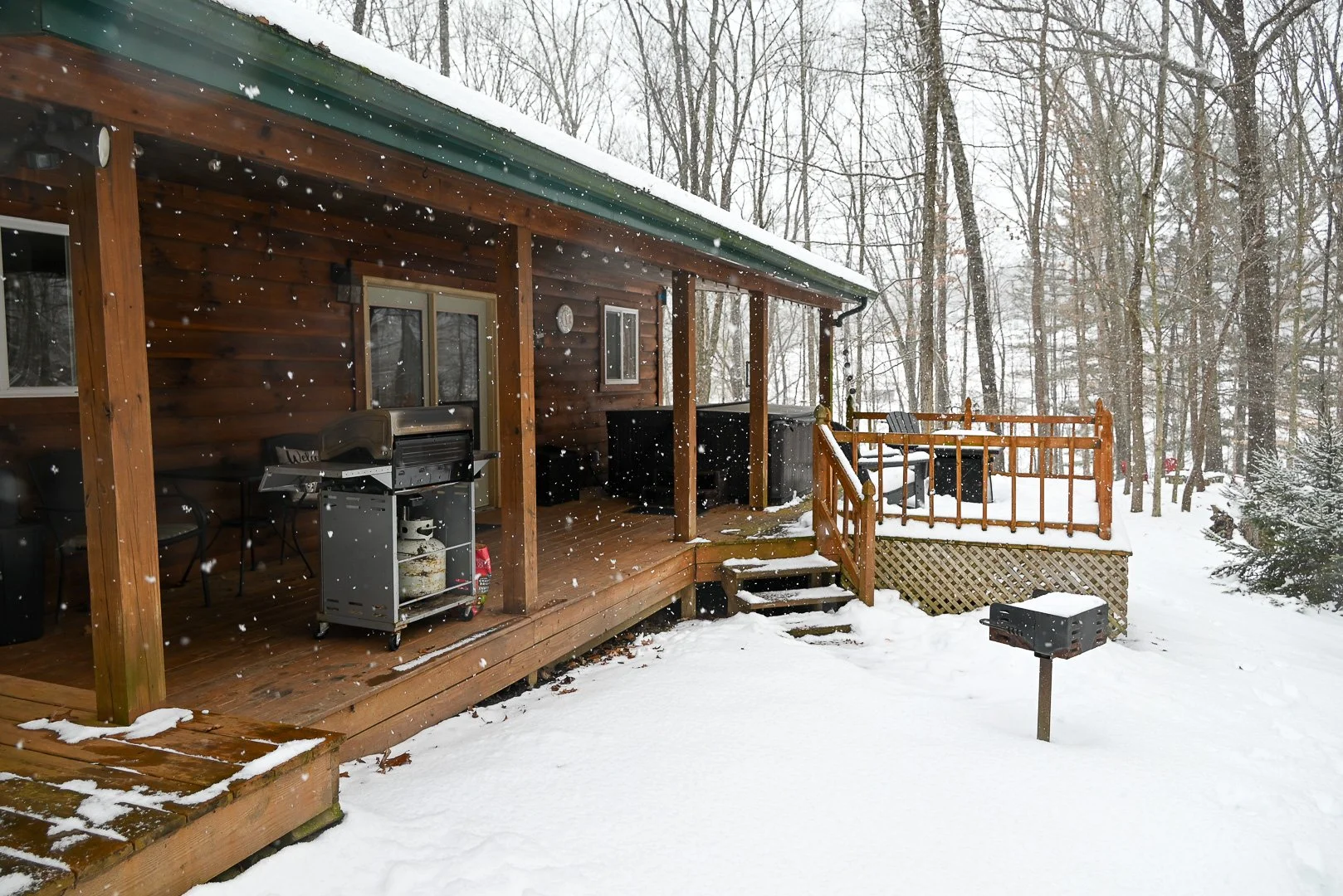 Snow-covered wooden cabin porch with a propane grill, sliding glass door, and adjacent deck, surrounded by leafless trees during active snowfall in a wooded winter setting.