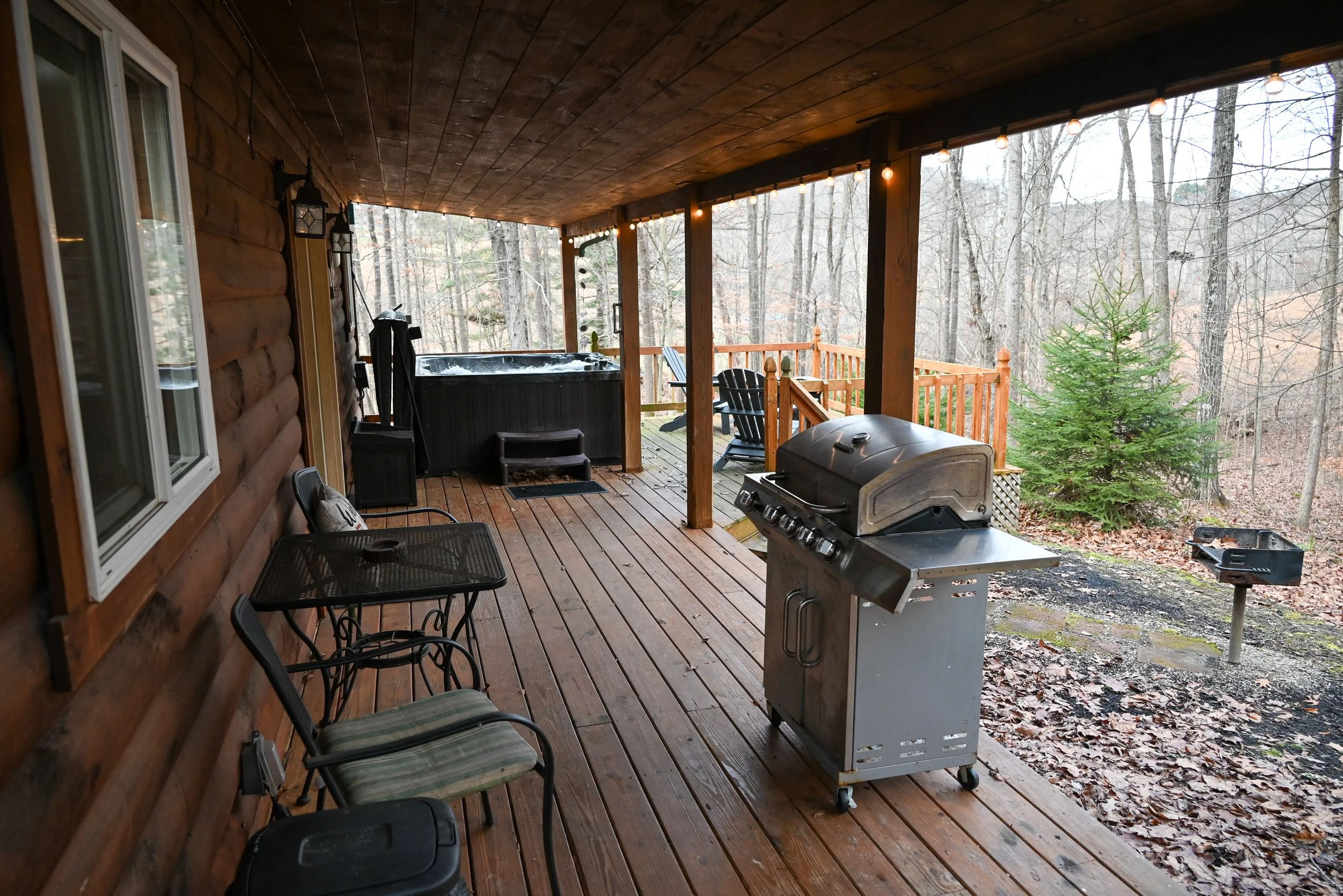 Covered porch at Beary Cozy Cabin in Hocking Hills featuring a gas grill, hot tub, outdoor seating, and wooded views for guests.