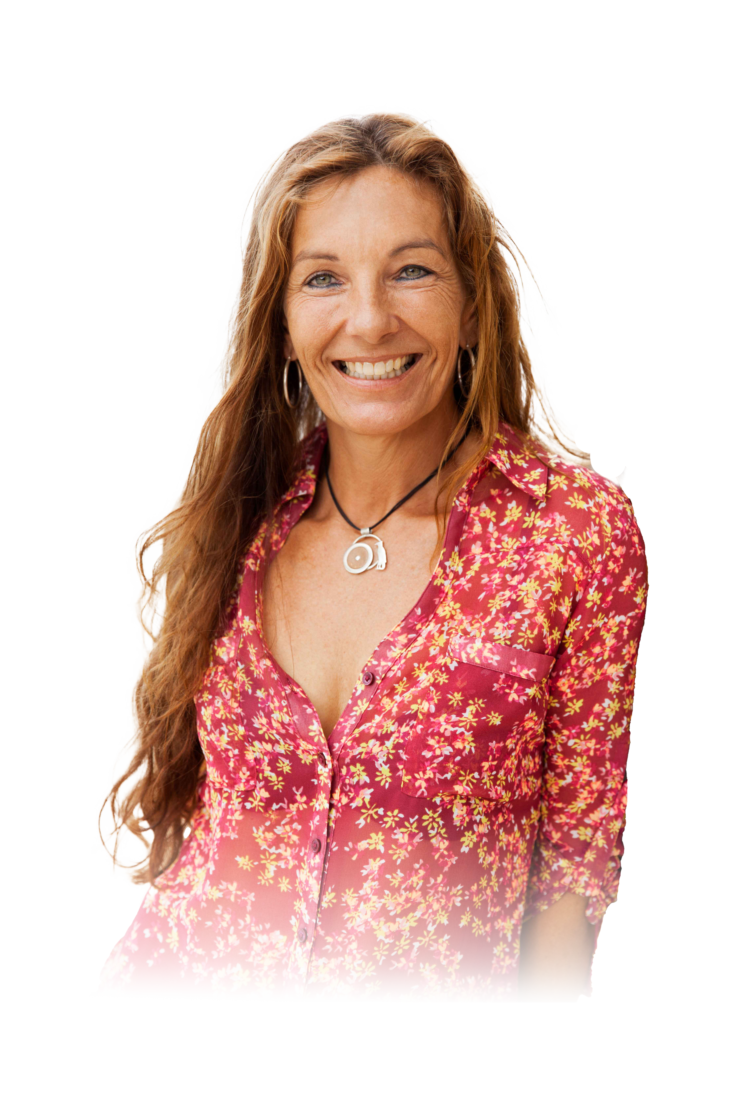 A woman with long red hair, smiling, wearing a red and yellow floral blouse, hoop earrings, and a black necklace with a circular pendant.