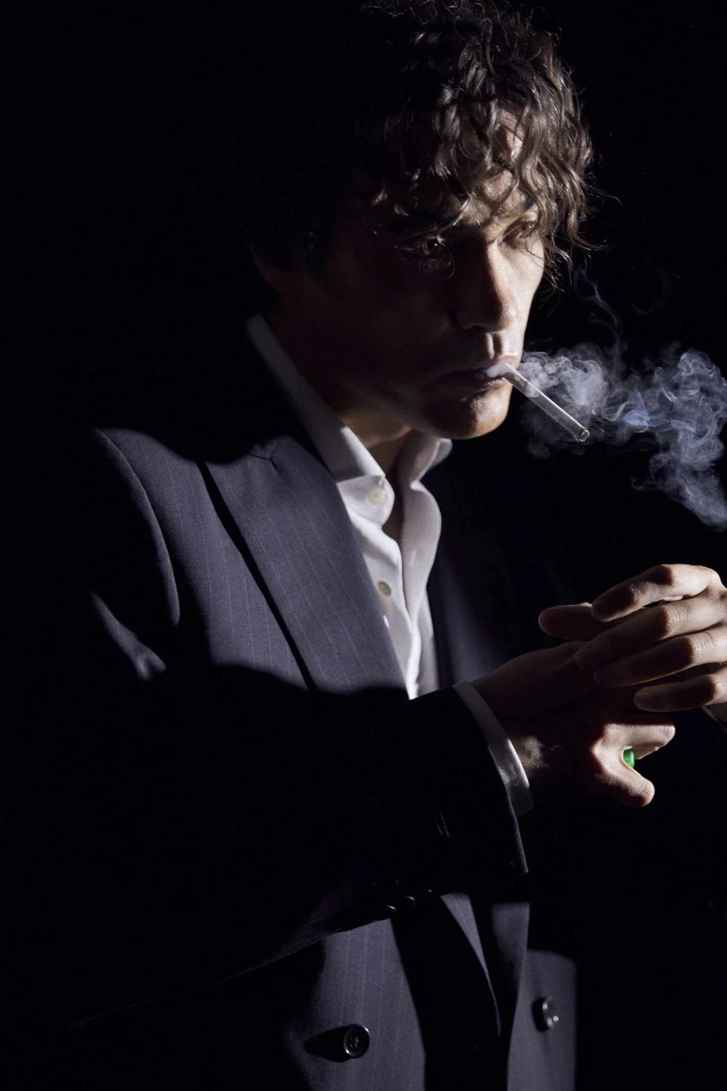 A man in a dark suit smoking a cigarette in a low-lit setting, with visible smoke and serious expression.
