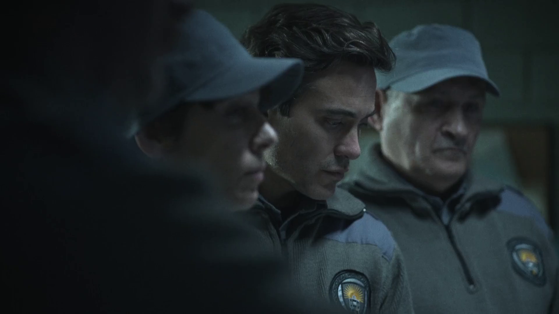 Three individuals in uniform seated close together, with serious expressions, likely in a somber or tense setting.